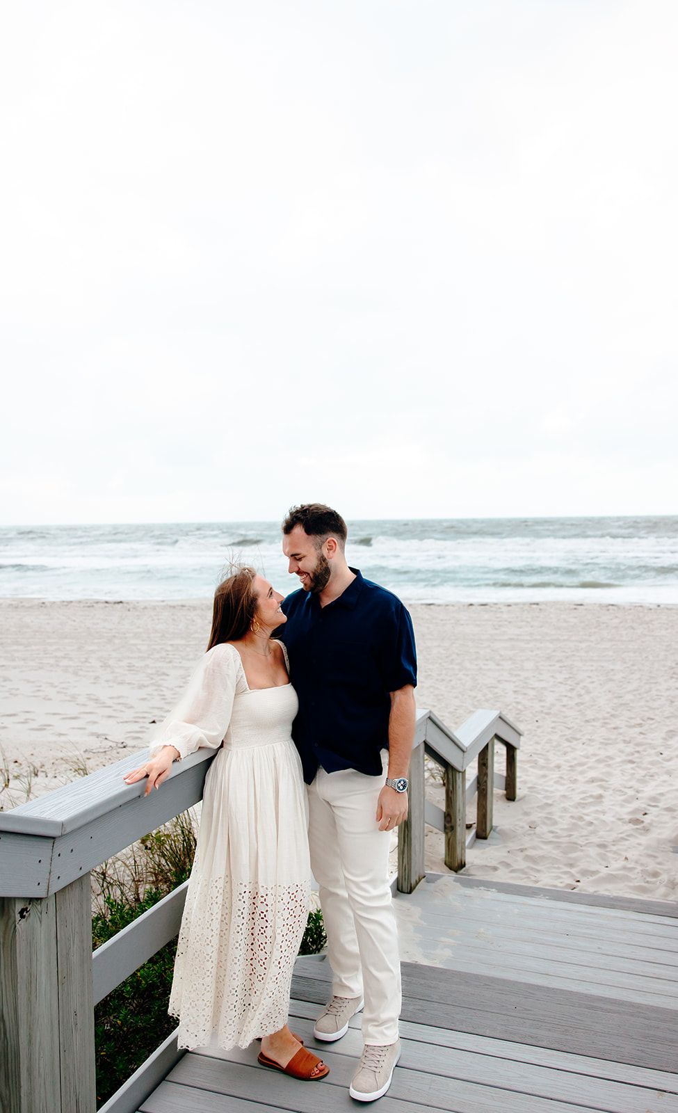 A couple smiles at each other on a beach boardwalk, woman in white dress, man in navy shirt, ocean backdrop.