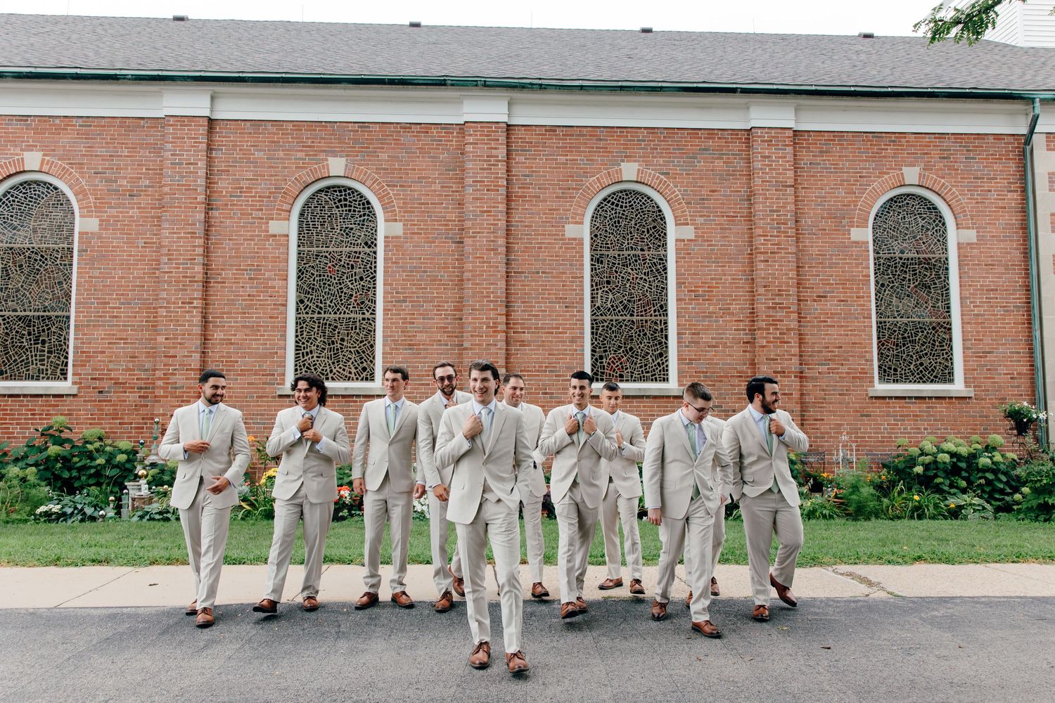 Groom and groomsmen in light beige suits posing outside a brick church on a sunny wedding day.