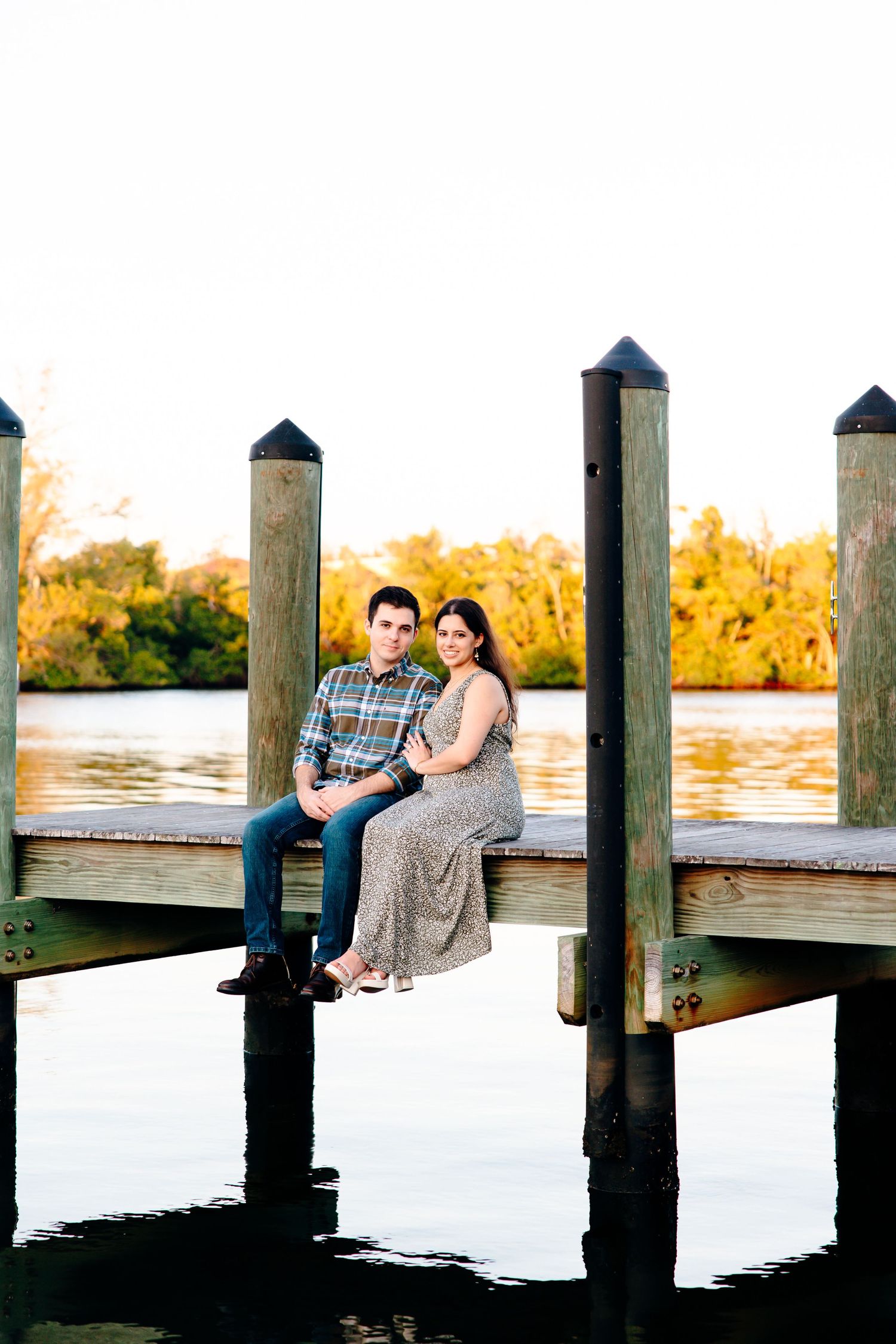 Couple sitting together on a wooden dock over calm water, surrounded by lush green trees at golden hour.