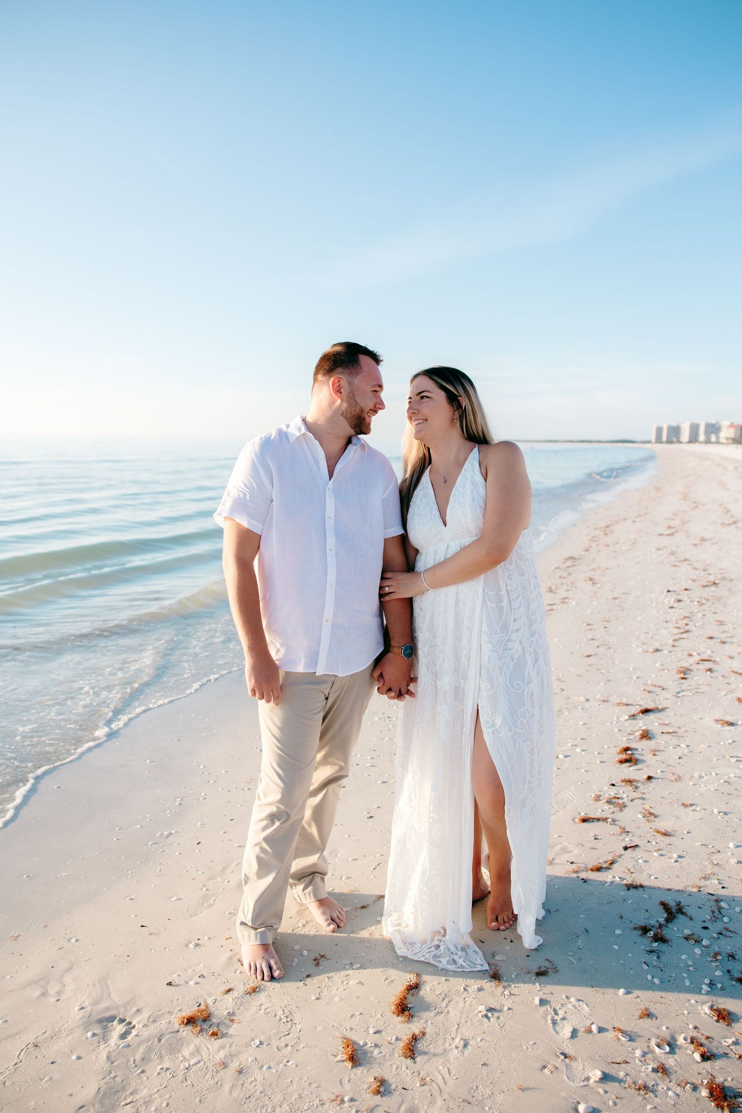 A couple walks hand in hand along a pristine white sandy beach with blue ocean waters and clear skies in the background.