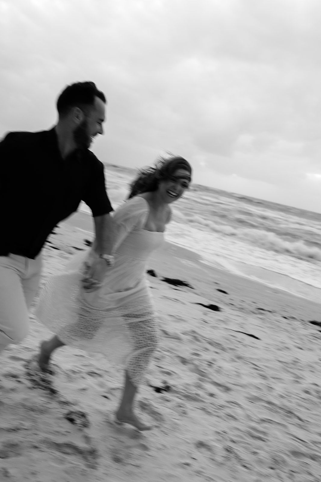 Black and white photo of a joyful couple running barefoot on a beach, woman in white dress, man in black shirt.