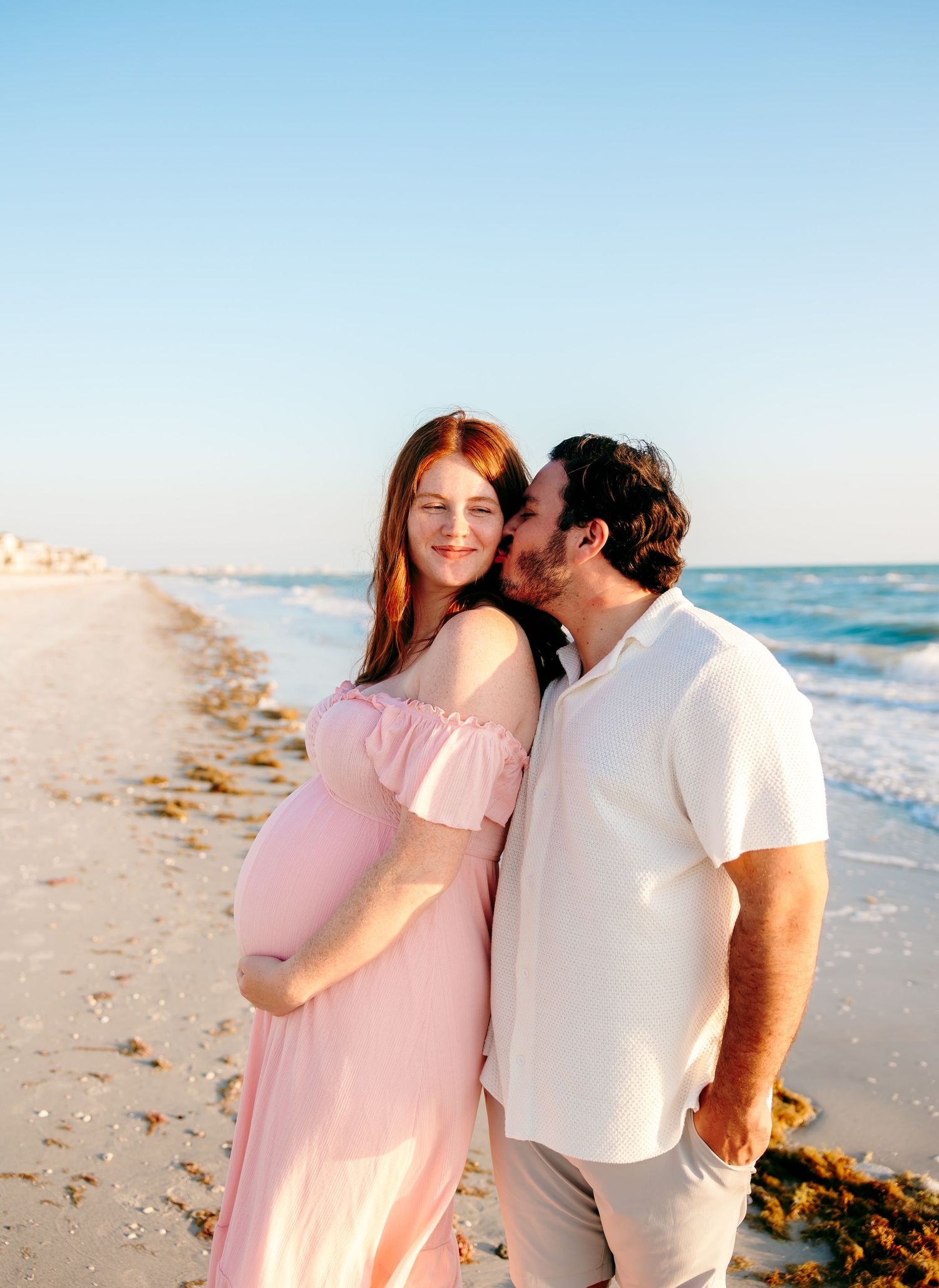 Pregnant woman in pink dress with partner kissing her cheek on a sunny beach during a maternity photo session.