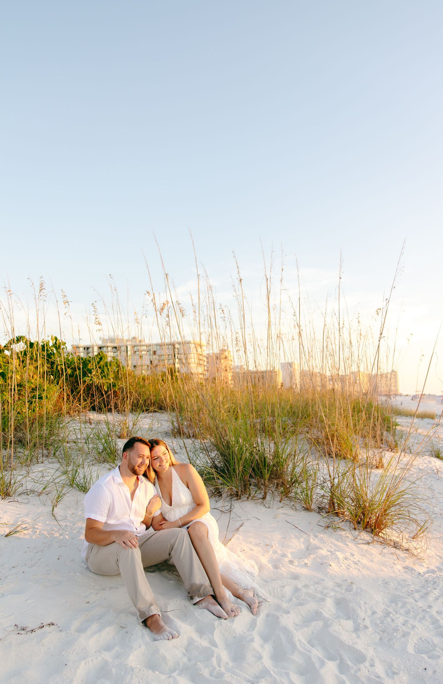 A couple sits together on white sand beach with sea oats and dunes during golden hour sunset.