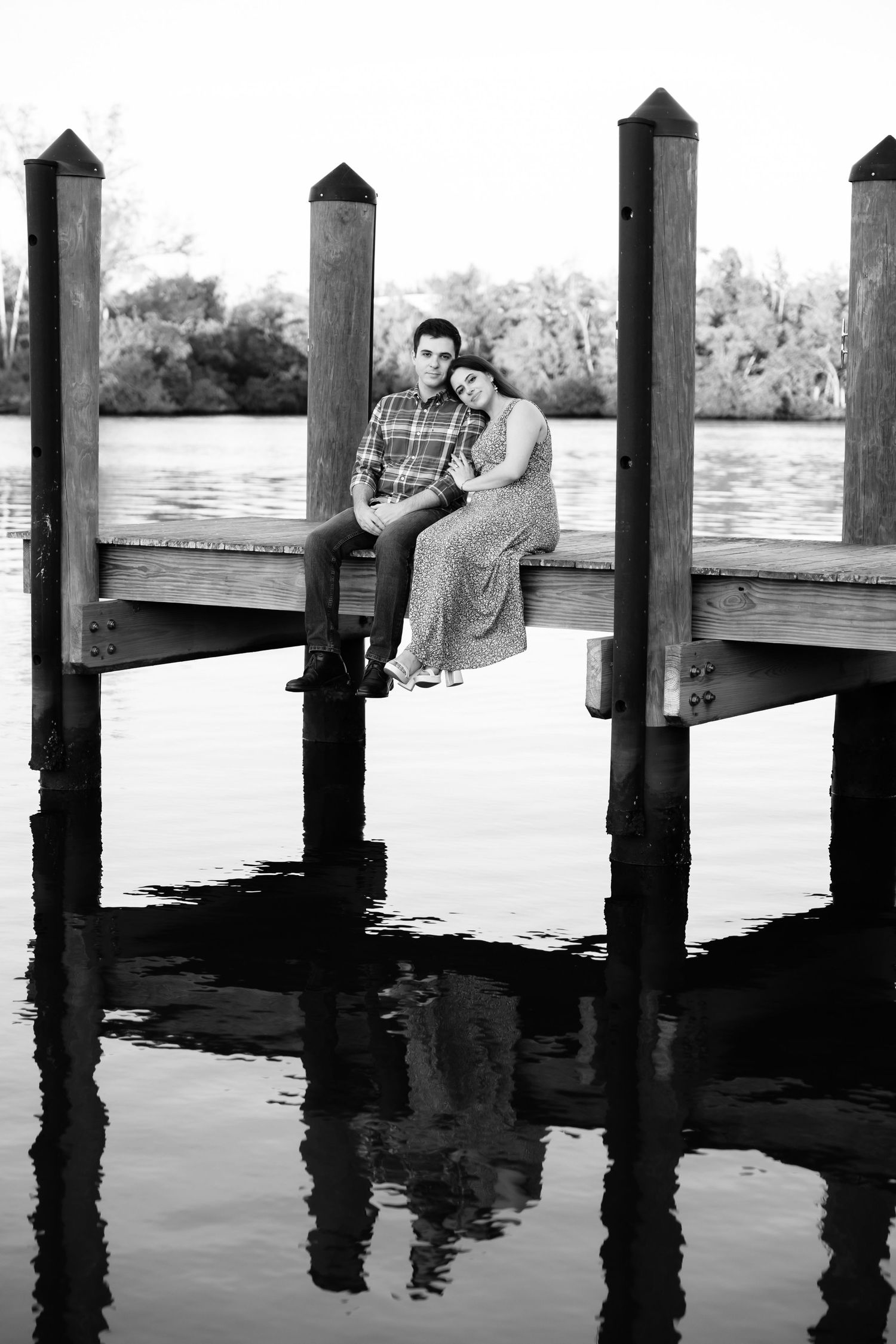 Couple sitting together on a wooden dock over calm water, black and white romantic engagement photo.