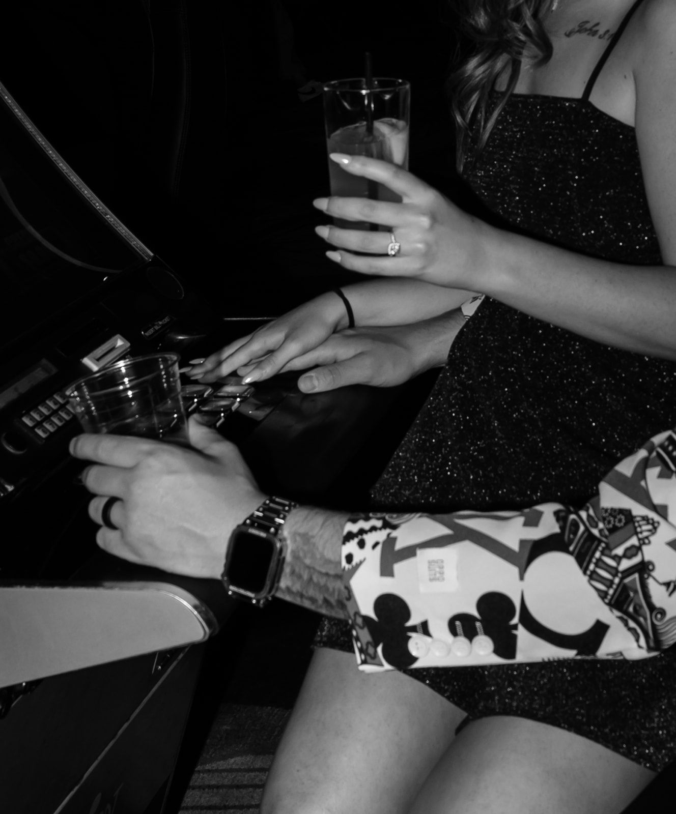 Black and white photo of a couple at a bar, woman in sparkly dress holding drink, man with tattoos wearing a watch.