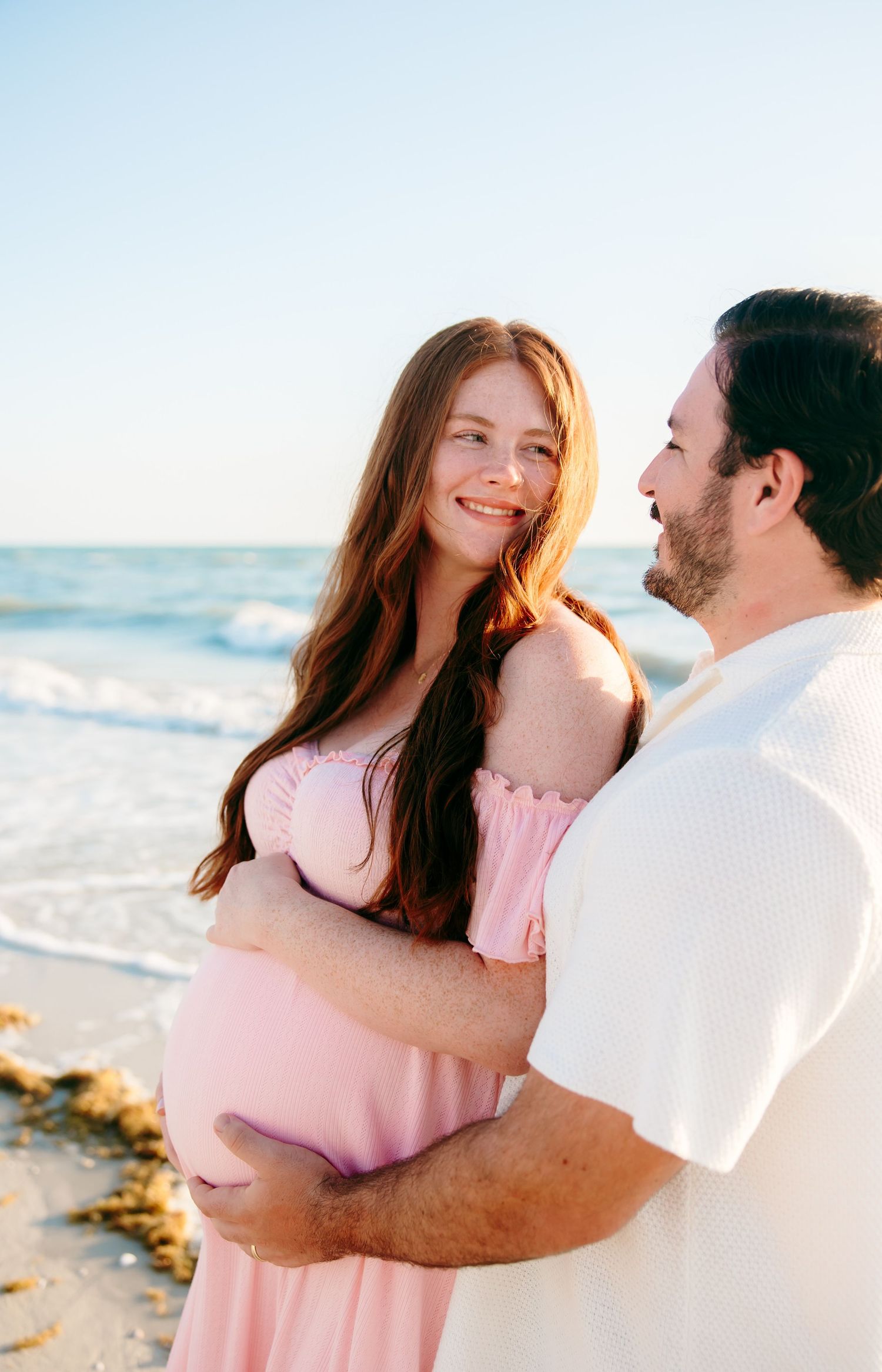 Pregnant woman in pink dress embraces partner on sunny beach, smiling lovingly during maternity photo shoot.