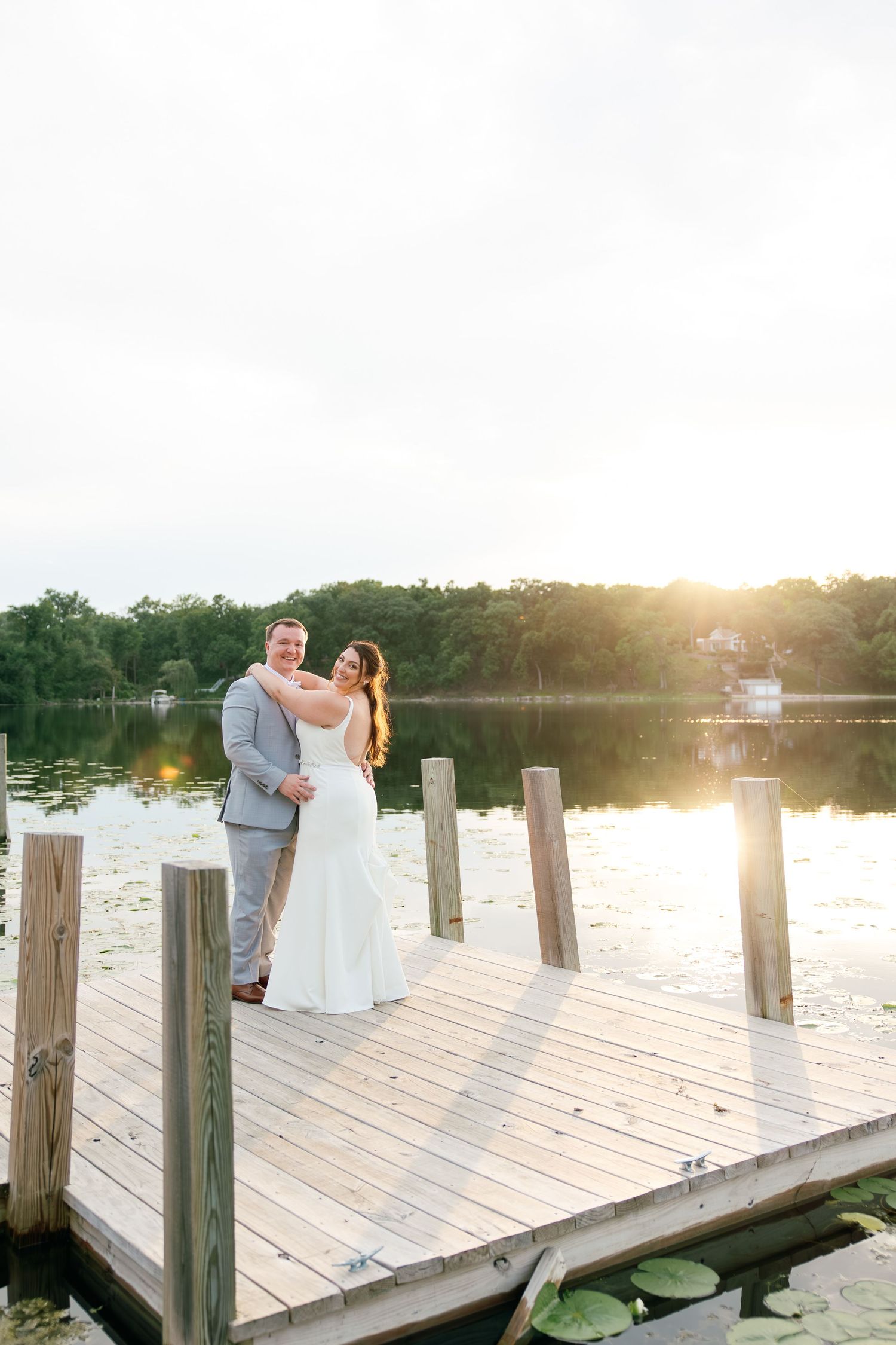 Bride and groom embrace on a sunlit wooden dock overlooking a serene lake at golden hour during their wedding.