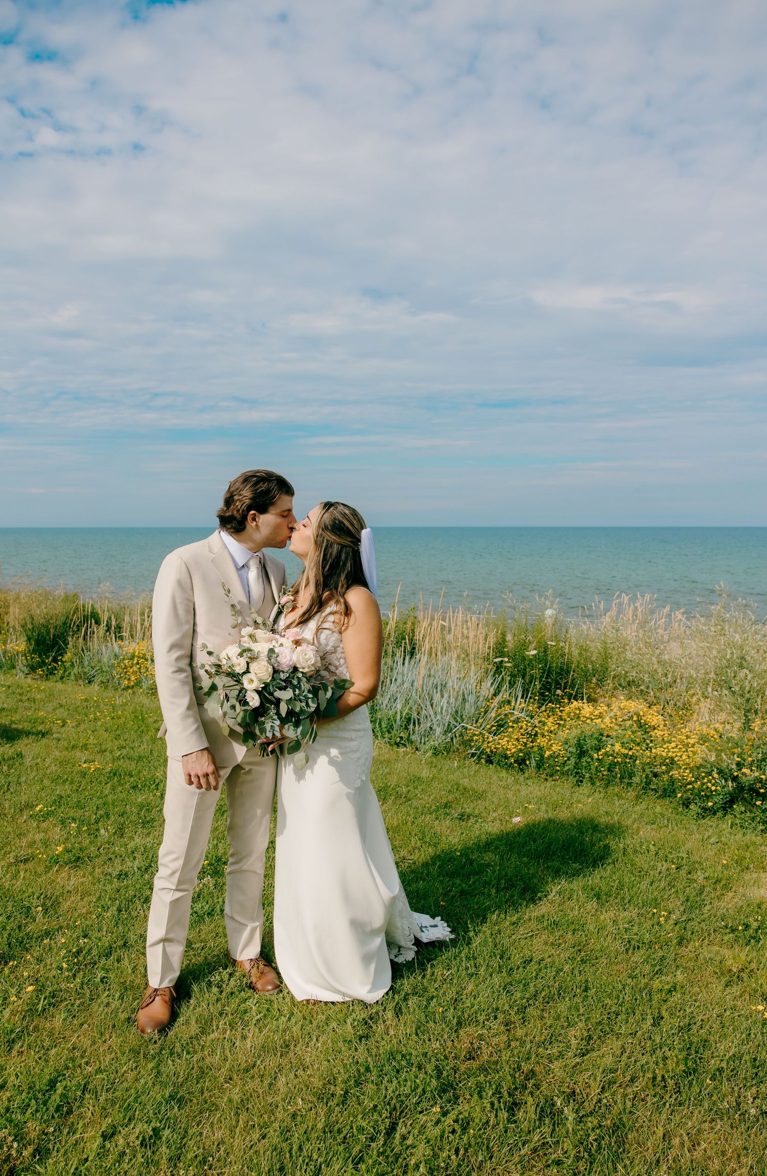 A bride and groom share a kiss on a grassy cliffside overlooking the ocean on their wedding day.