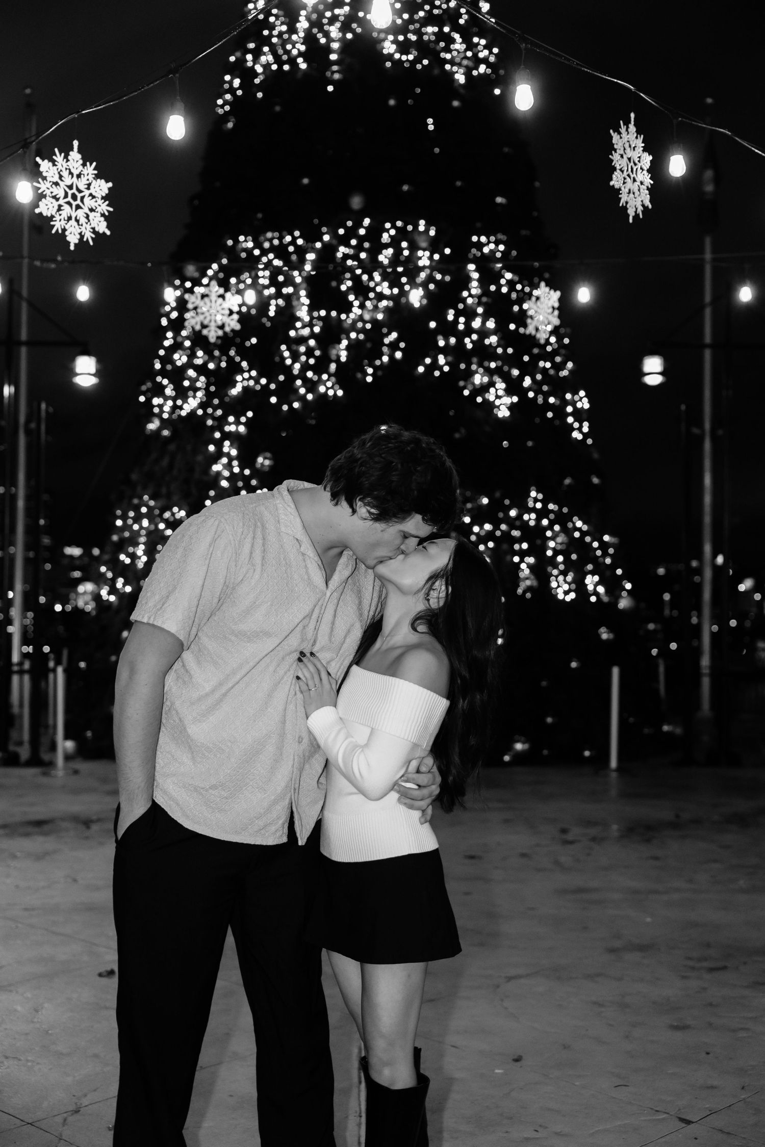Black and white photo of a couple kissing in front of a lit Christmas tree with snowflake decorations at night.