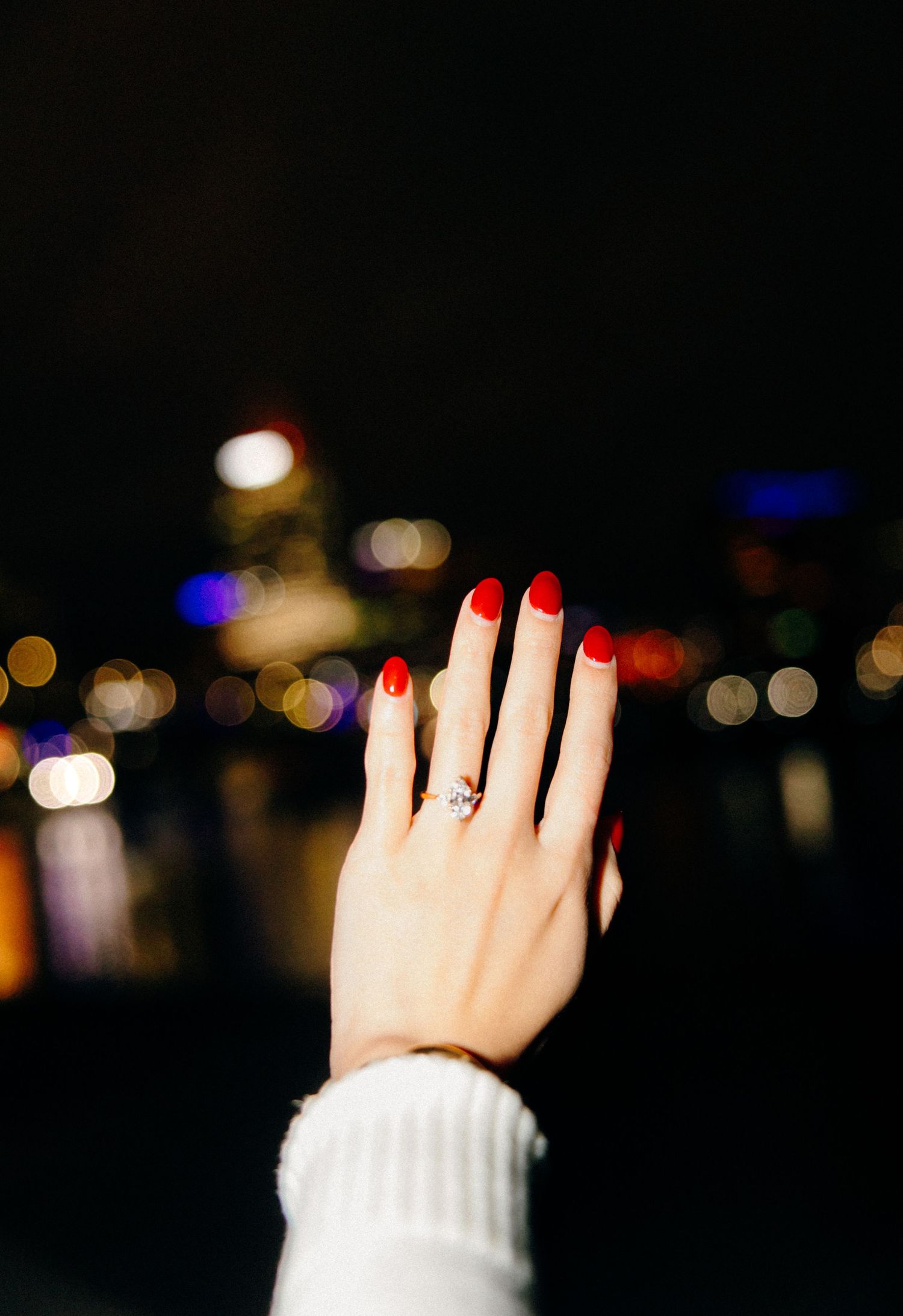 Woman displaying engagement ring with red nails against bokeh city lights at night.