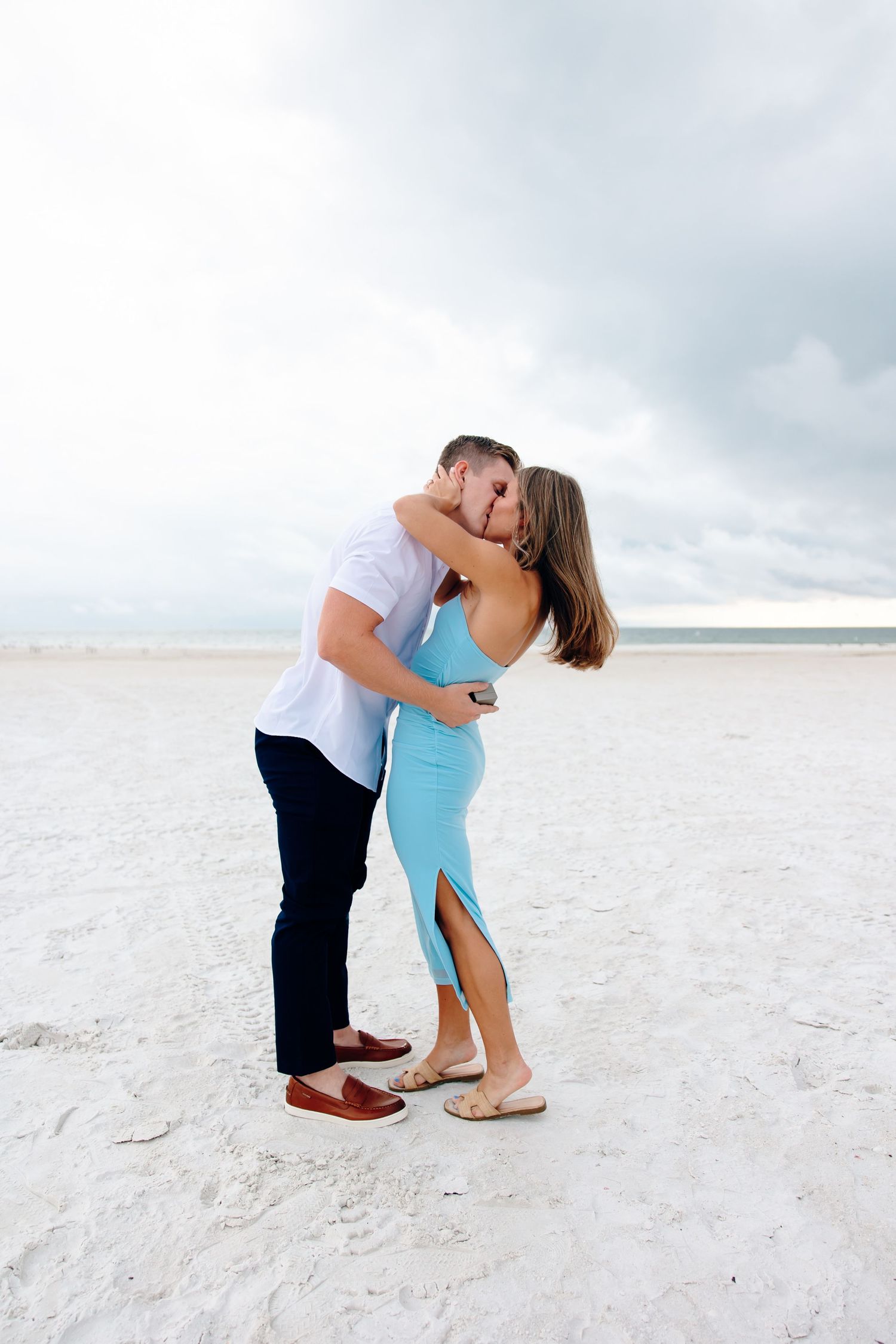 Couple kissing on a white sand beach, woman in turquoise dress, man in white shirt and dark pants.