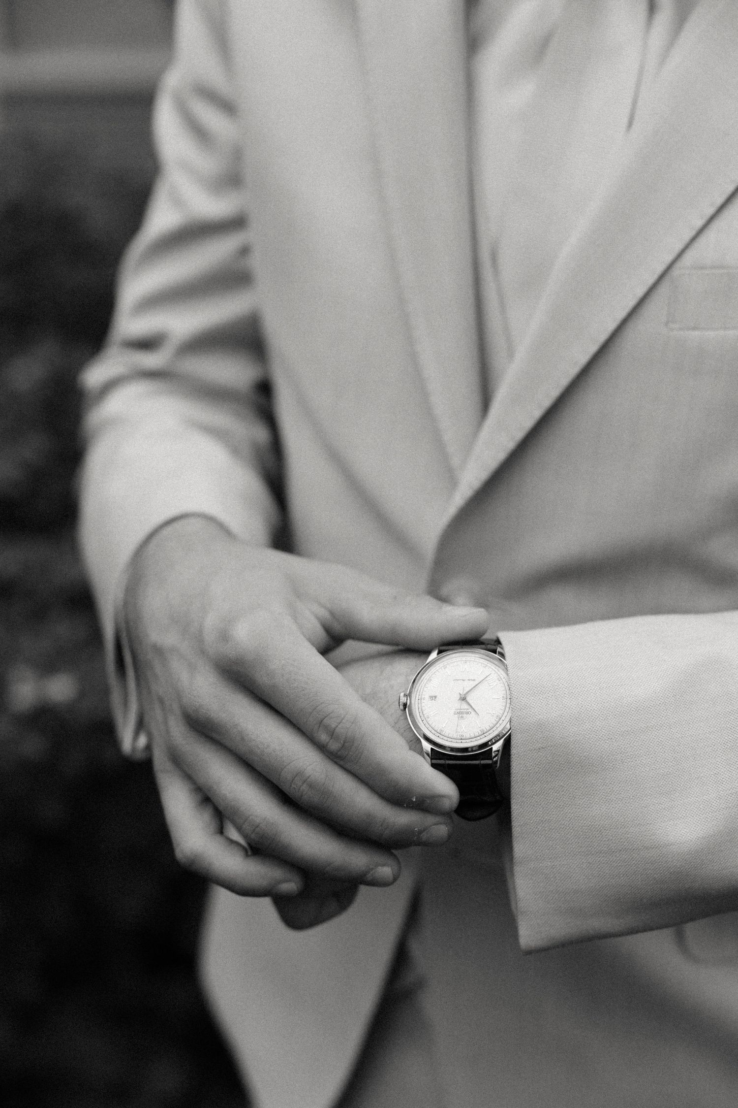 Black and white photo of a groom in a light suit adjusting his classic analog wristwatch, with a wedding ring visible.