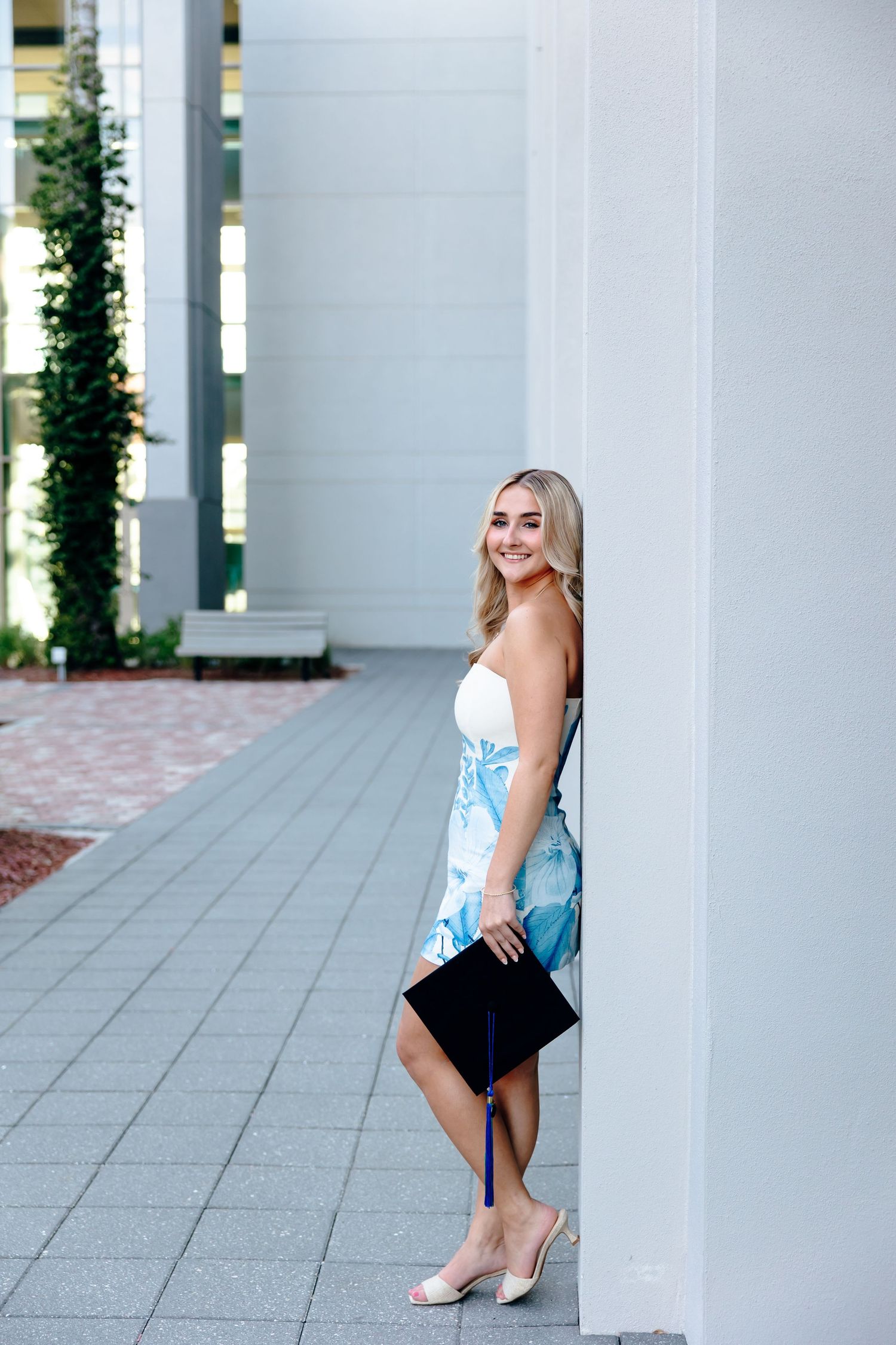 Young blonde woman in floral strapless dress holds graduation cap, smiling beside white column outdoors.