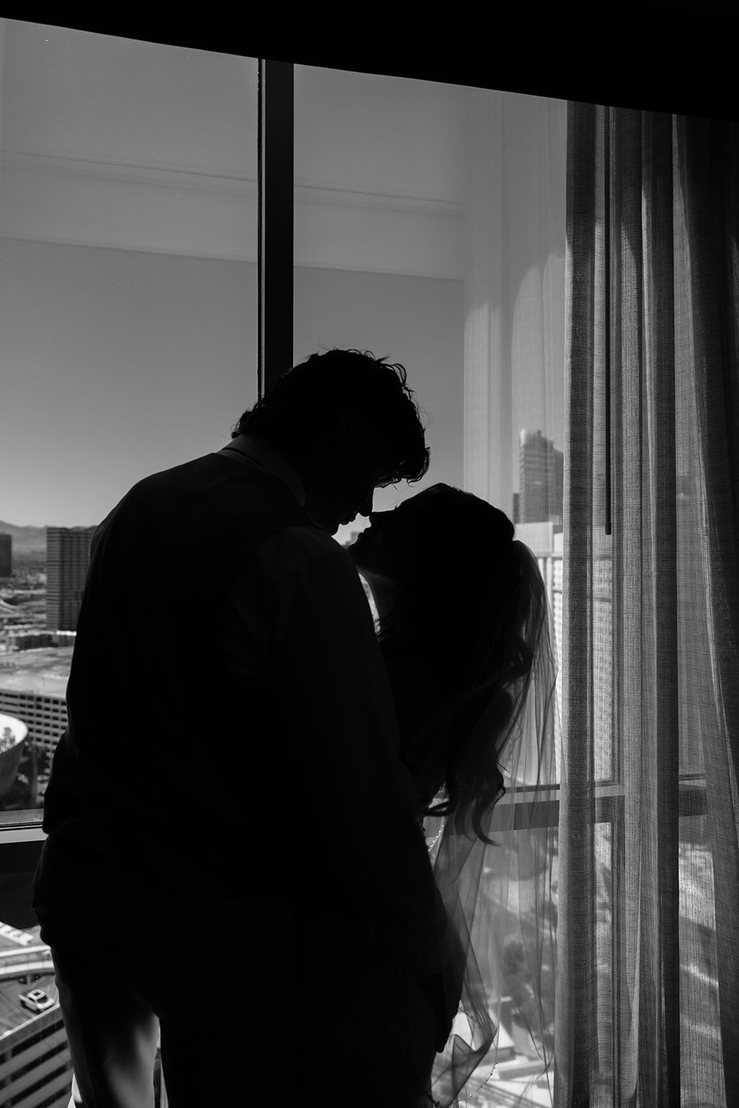 Silhouette of bride and groom sharing a kiss by a sunlit window with city views, black and white.