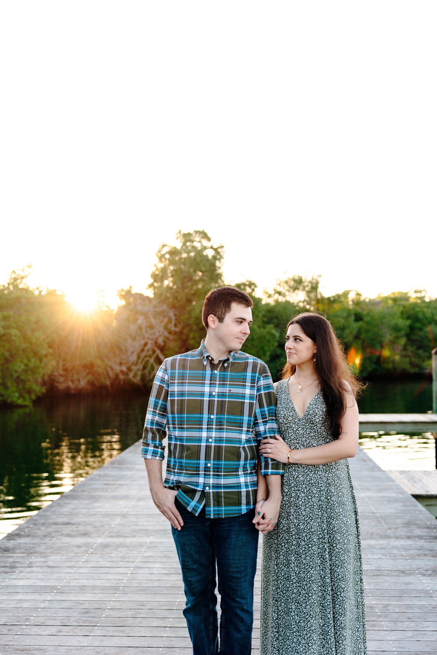Couple holding hands on a wooden dock at sunset with a serene lake and lush greenery in the background.