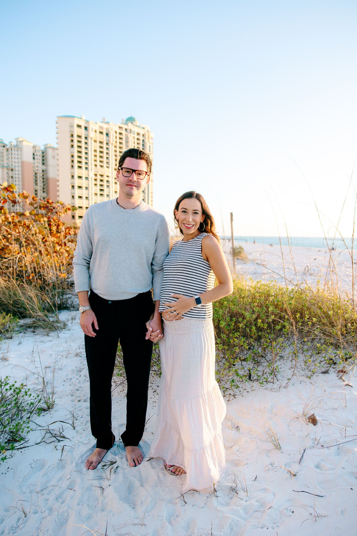 Couple posing on sandy beach with coastal buildings behind them, woman in striped top holding baby bump.