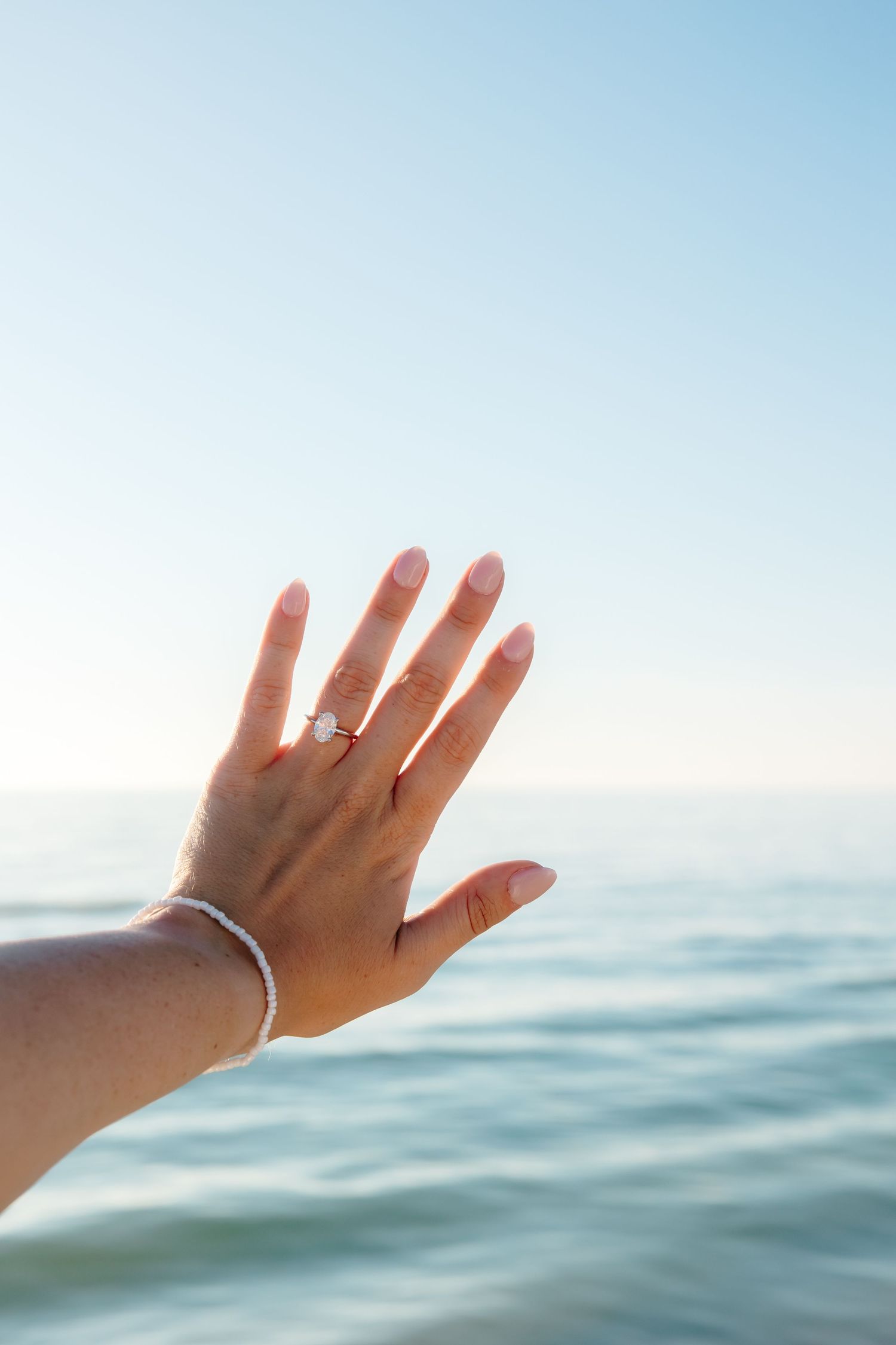 Hand wearing a ring and bracelet reaches toward the ocean horizon against a bright blue sky.