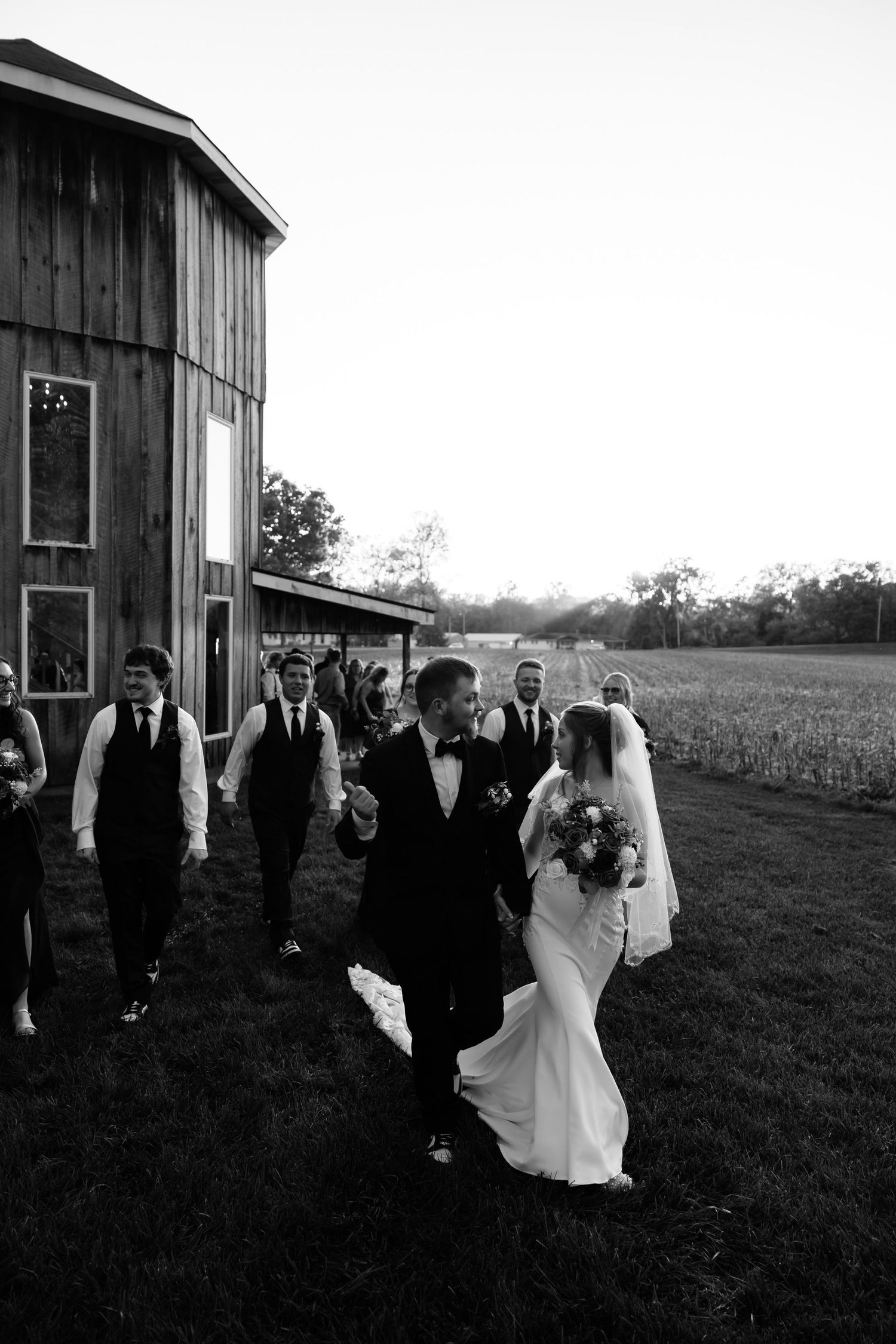 Black and white photo of bride and groom walking with wedding party beside a rustic barn on a farm.
