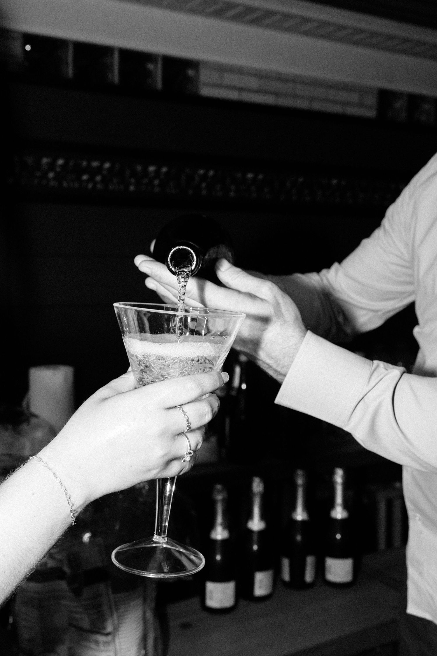 Black and white photo of bartender pouring champagne into a martini glass held by a guest at a bar.