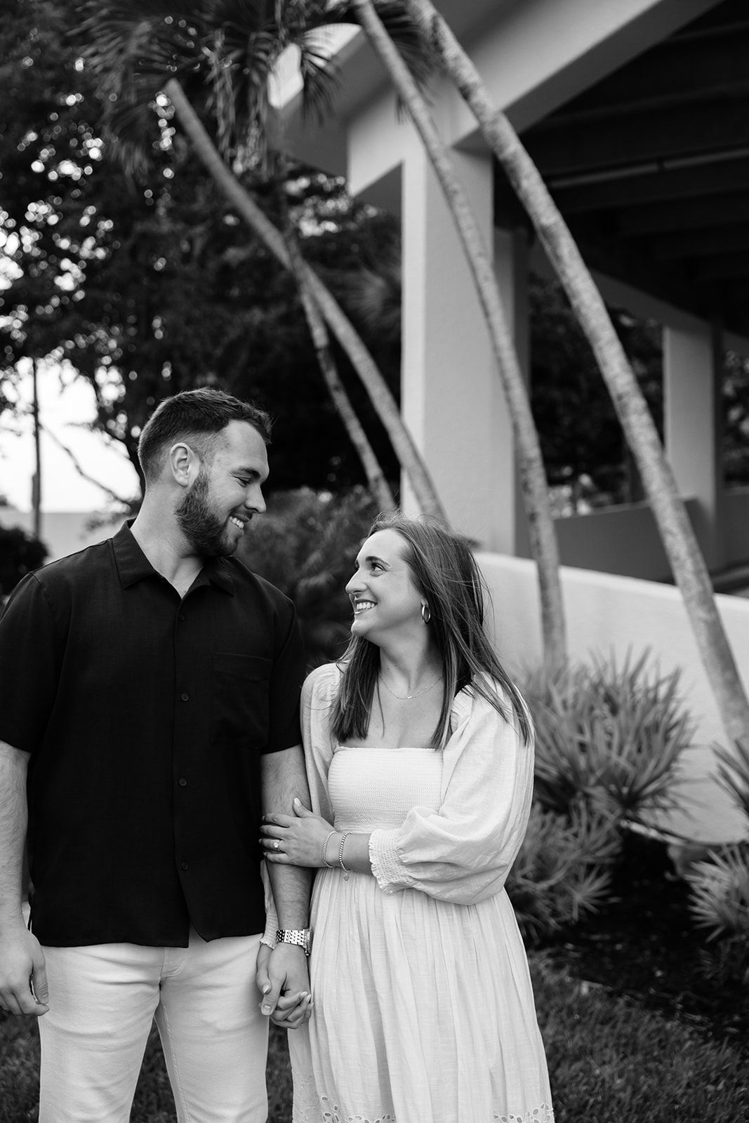 Black and white photo of smiling couple holding hands outdoors near palm trees and modern building.
