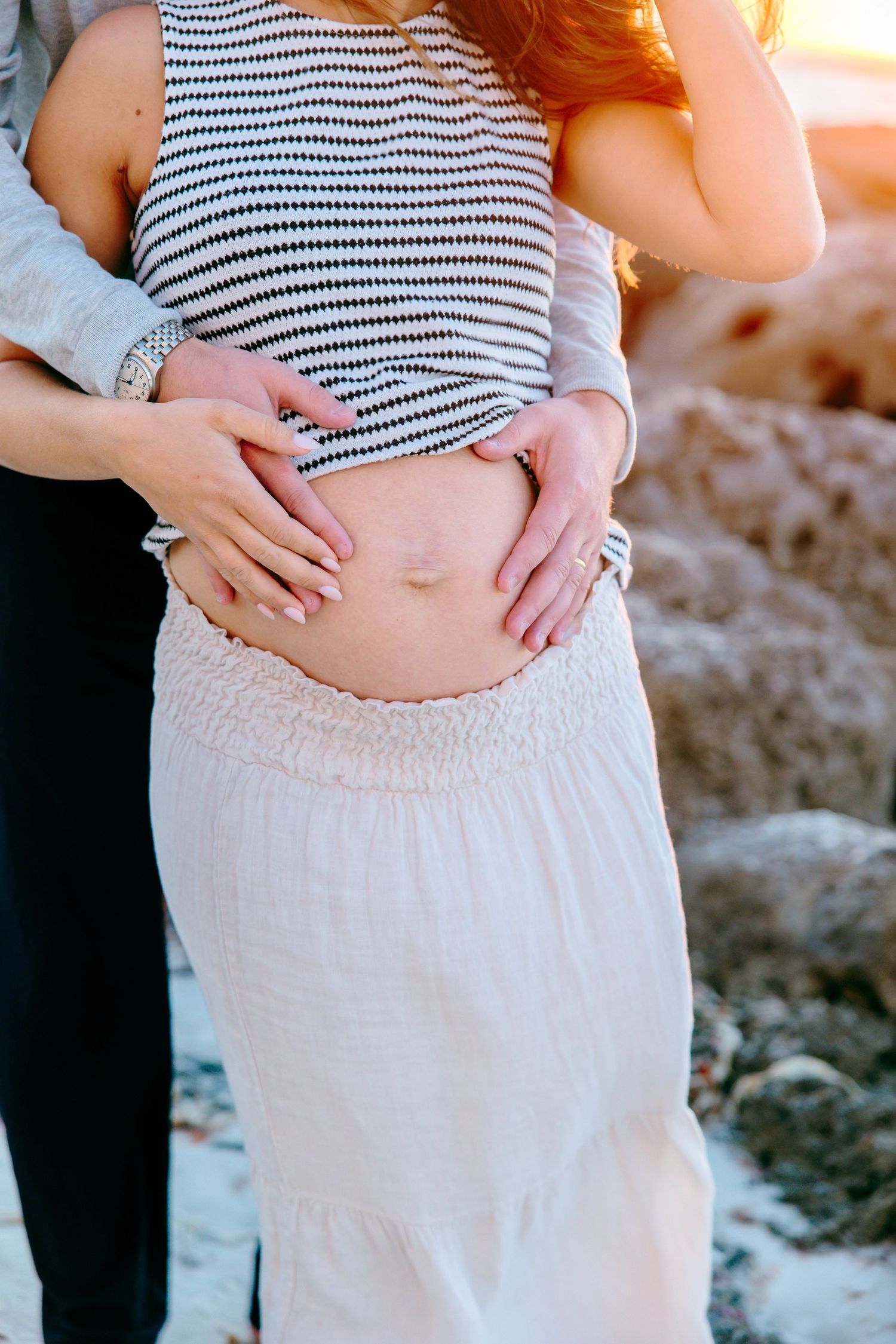 Pregnant woman in striped top and white skirt with partner's hands forming heart shape on baby bump at beach.