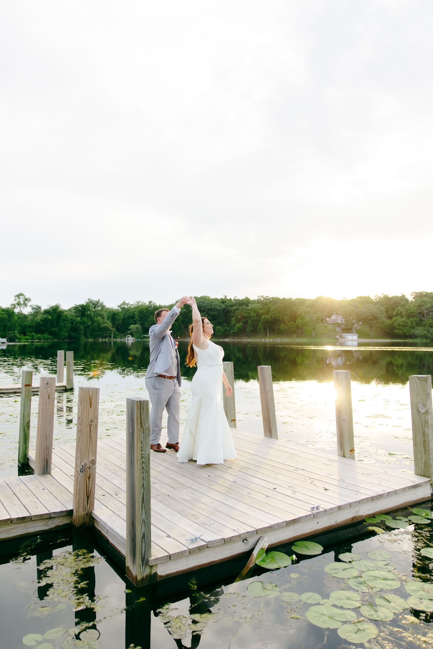 Bride and groom dance on a wooden dock at sunset, surrounded by a serene lily pad lake.