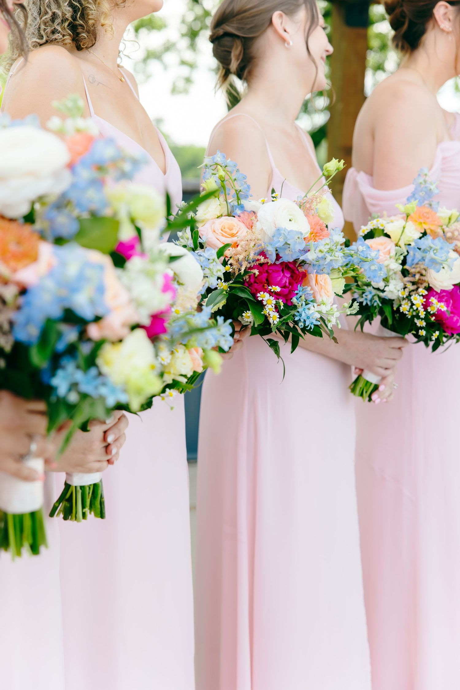 Bridesmaids in blush pink gowns holding colorful wedding bouquets with blue, pink, and white flowers.