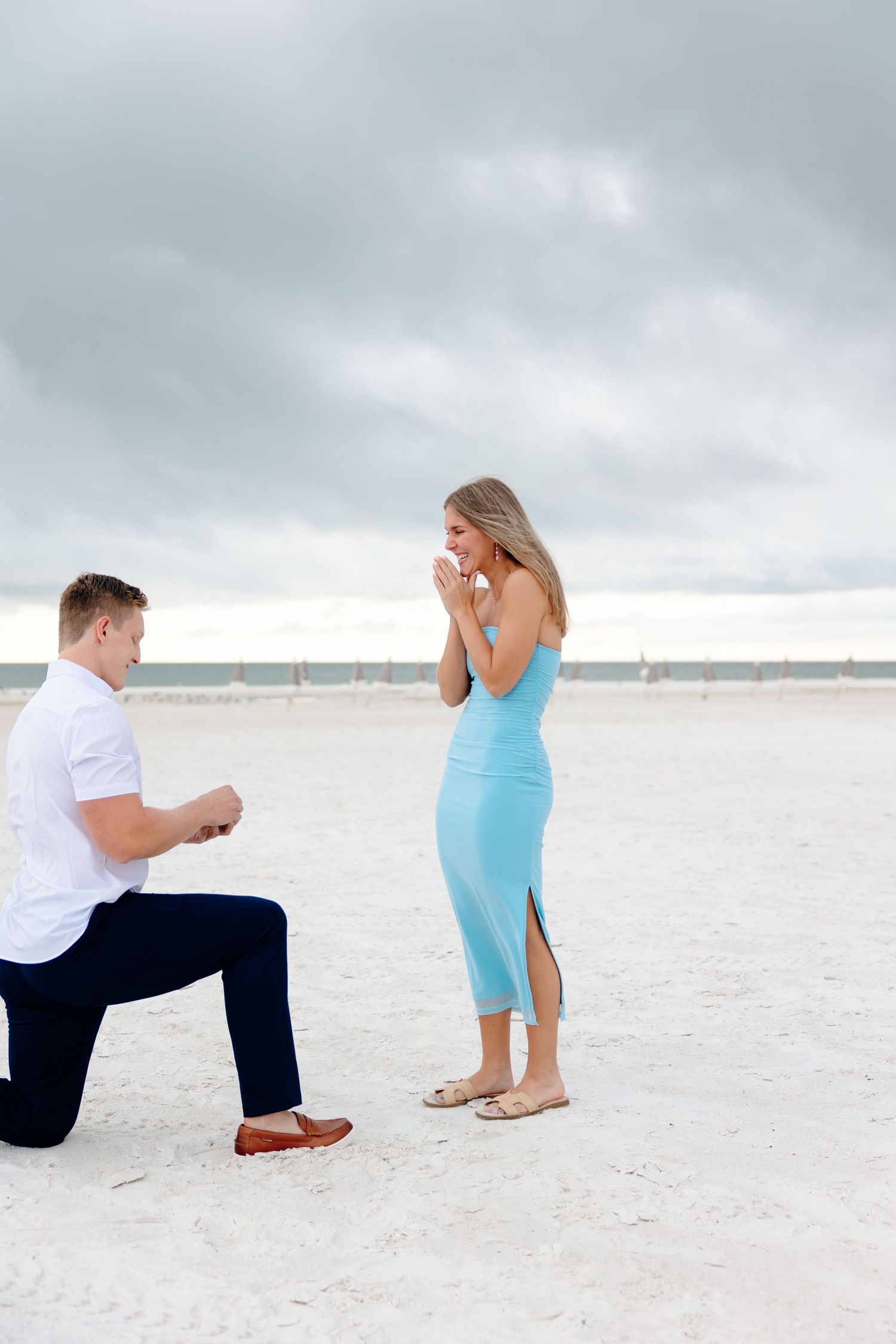 Man proposing on one knee to woman in blue dress on sandy beach with cloudy sky backdrop.