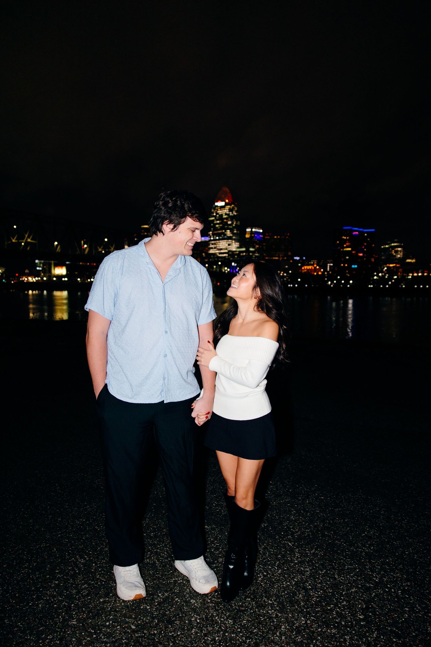 Couple holding hands smiling at each other at night with city skyline lights reflecting on water behind them.