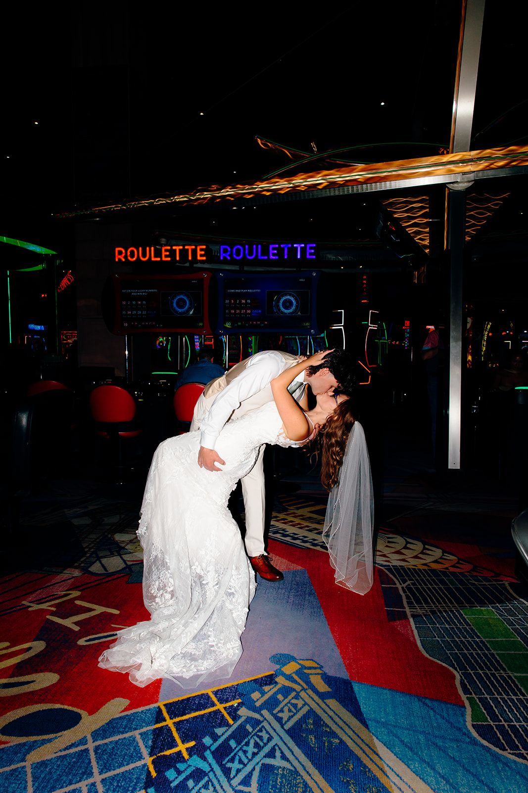 Bride and groom share a romantic dip kiss on a colorful dance floor near roulette signs at a casino wedding venue.