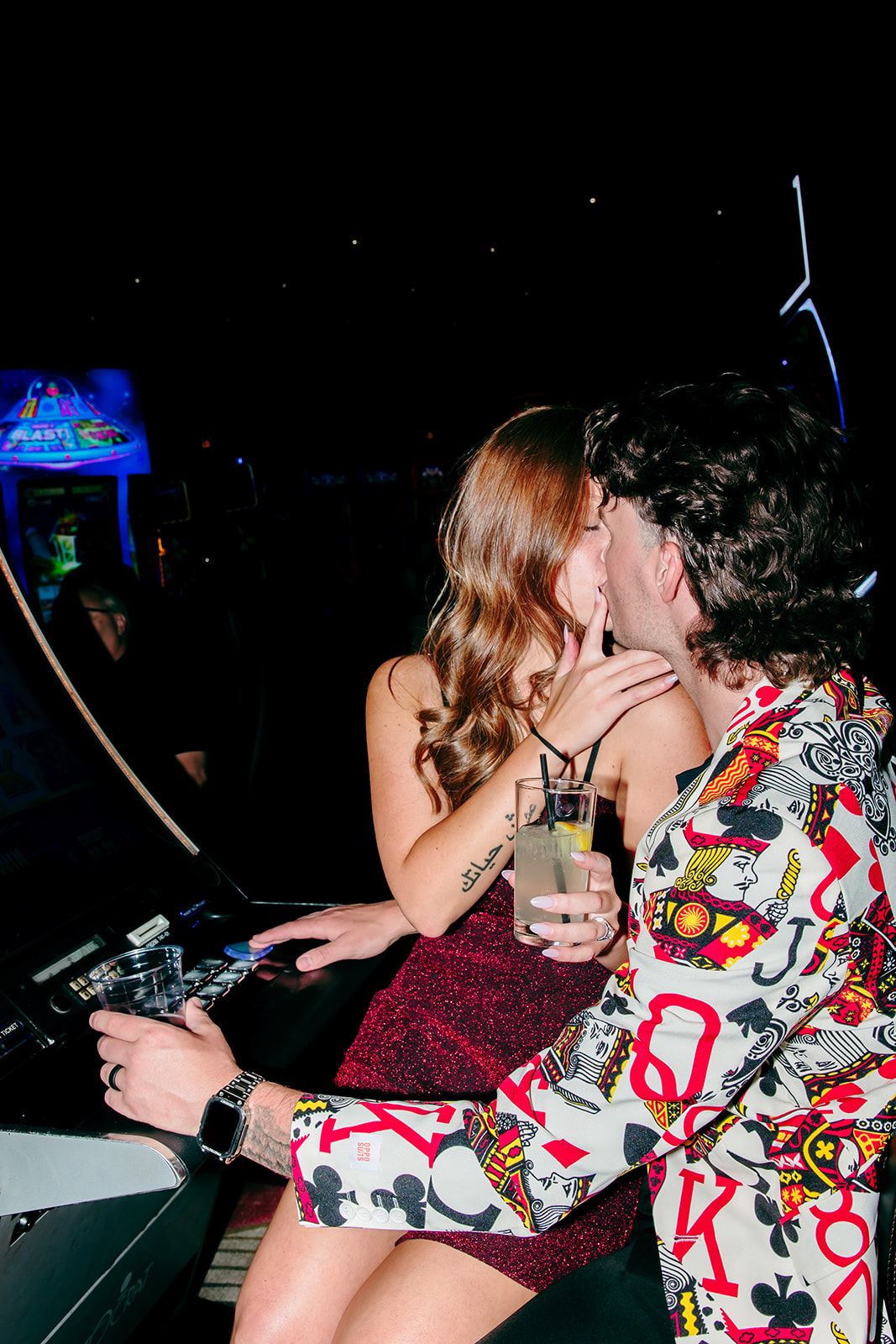 Couple kissing at arcade bar, man in playing card suit holding drink, woman in red dress with tattoo.