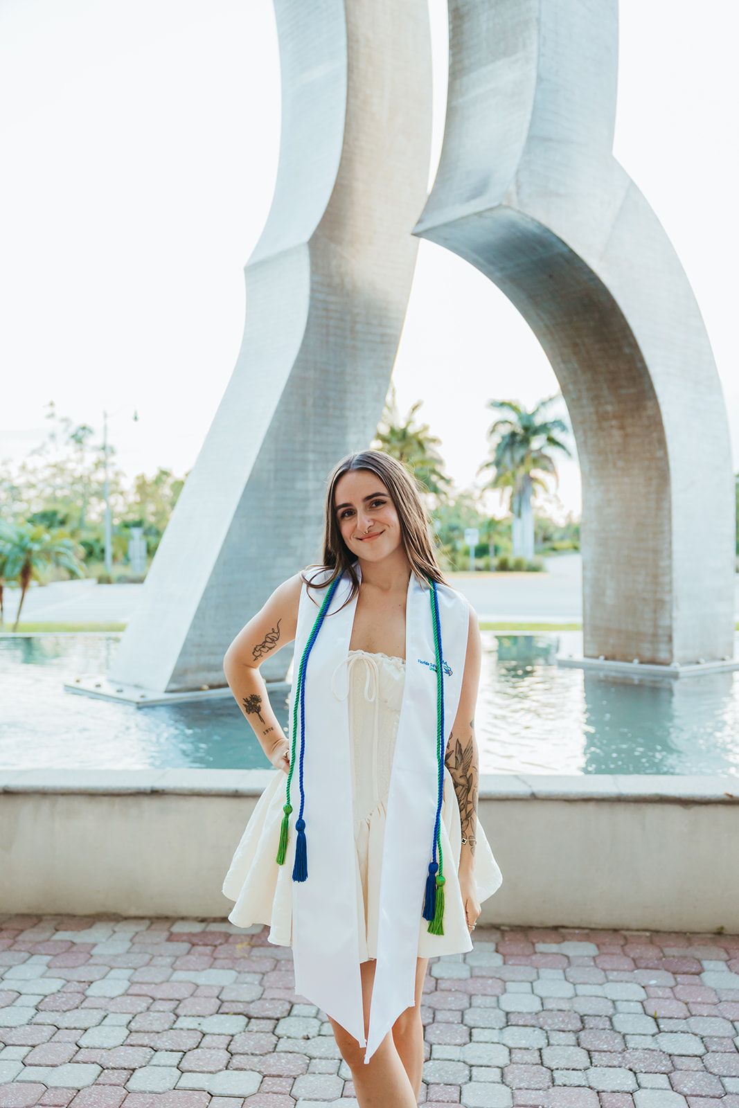 Graduate in white dress and blue stole poses near architectural columns by waterfront.