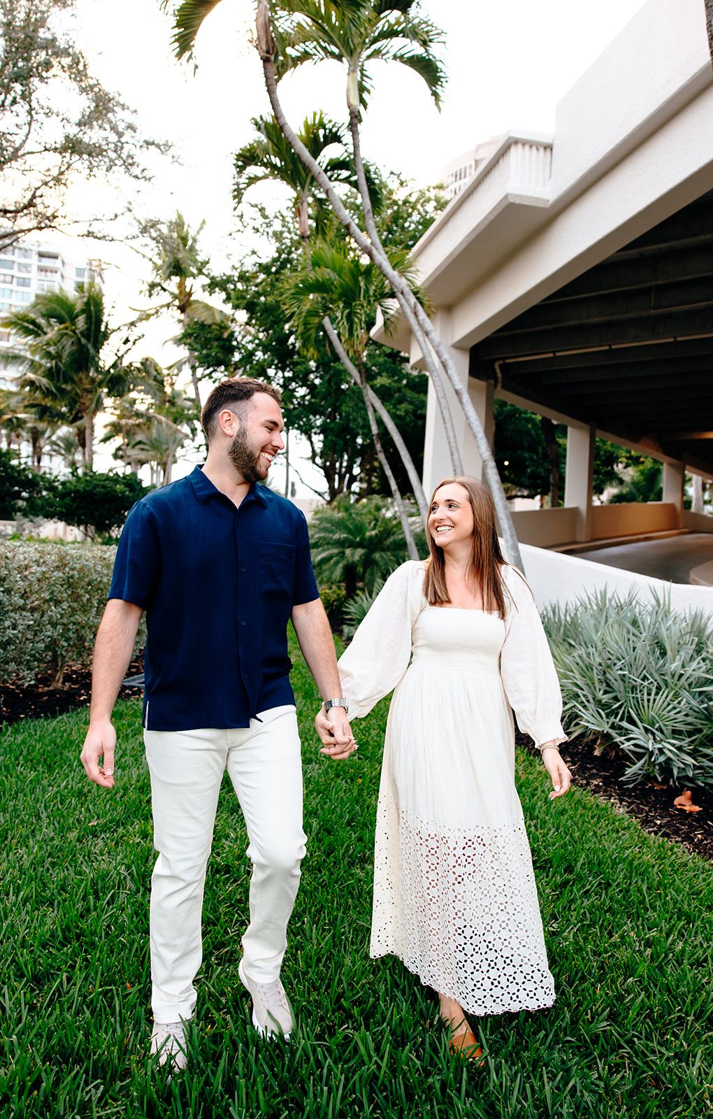 Couple holding hands walking on green grass near palm trees, man in navy shirt and woman in white lace dress.