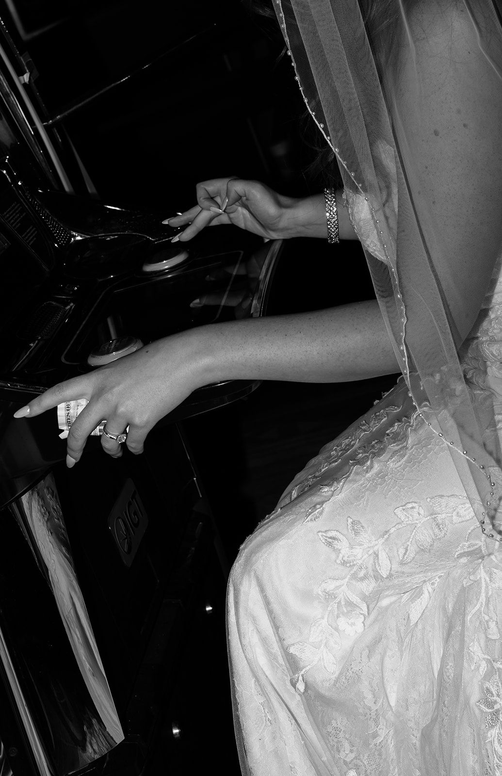 Black and white photo of a bride in lace gown with rings and bracelet, reaching out from a vehicle door.