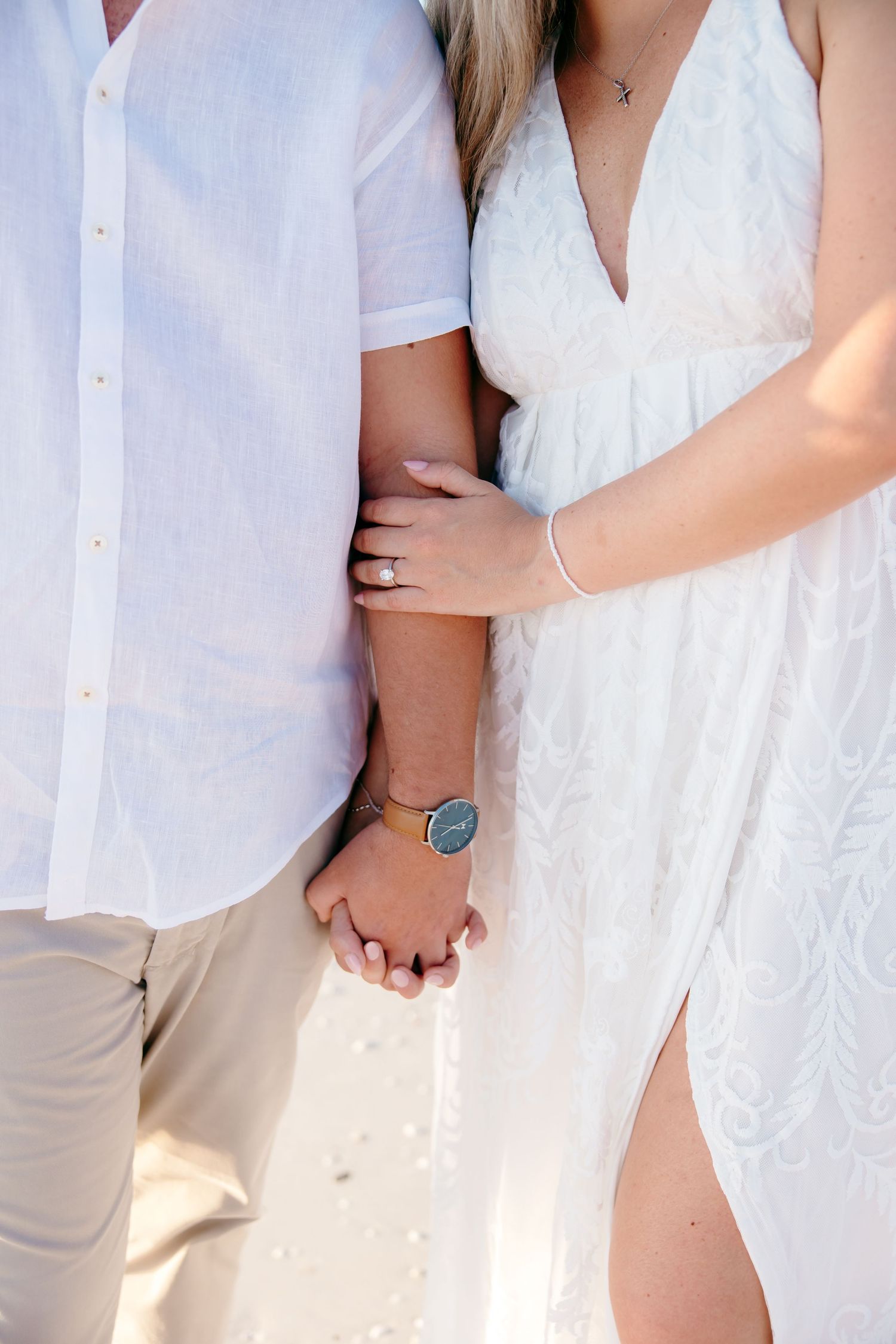 Close-up of couple holding hands, wearing white summer clothing with a turquoise bracelet visible.