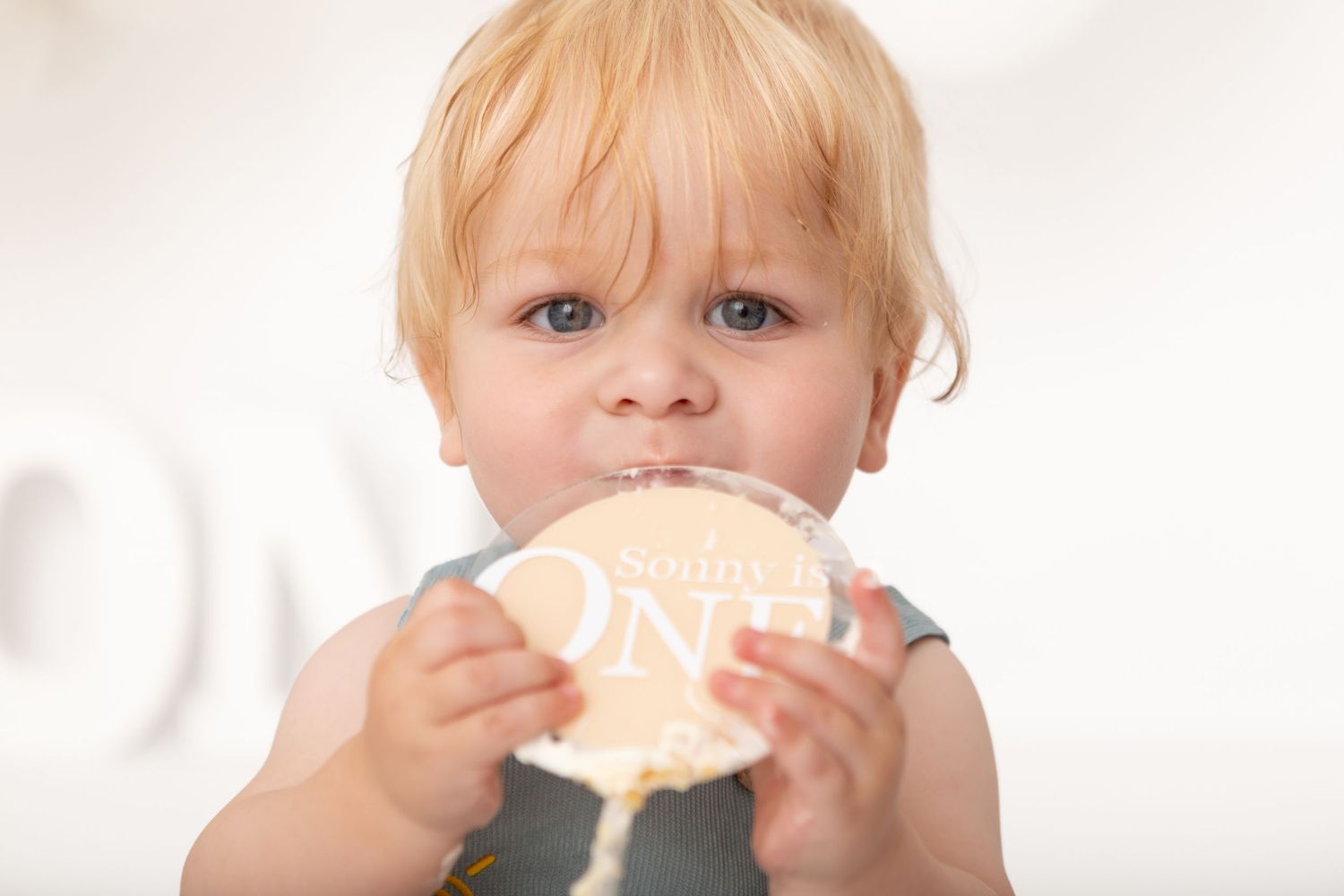 Blonde toddler chewing on a wooden milestone disc sign reading ONE, celebrating their first birthday.