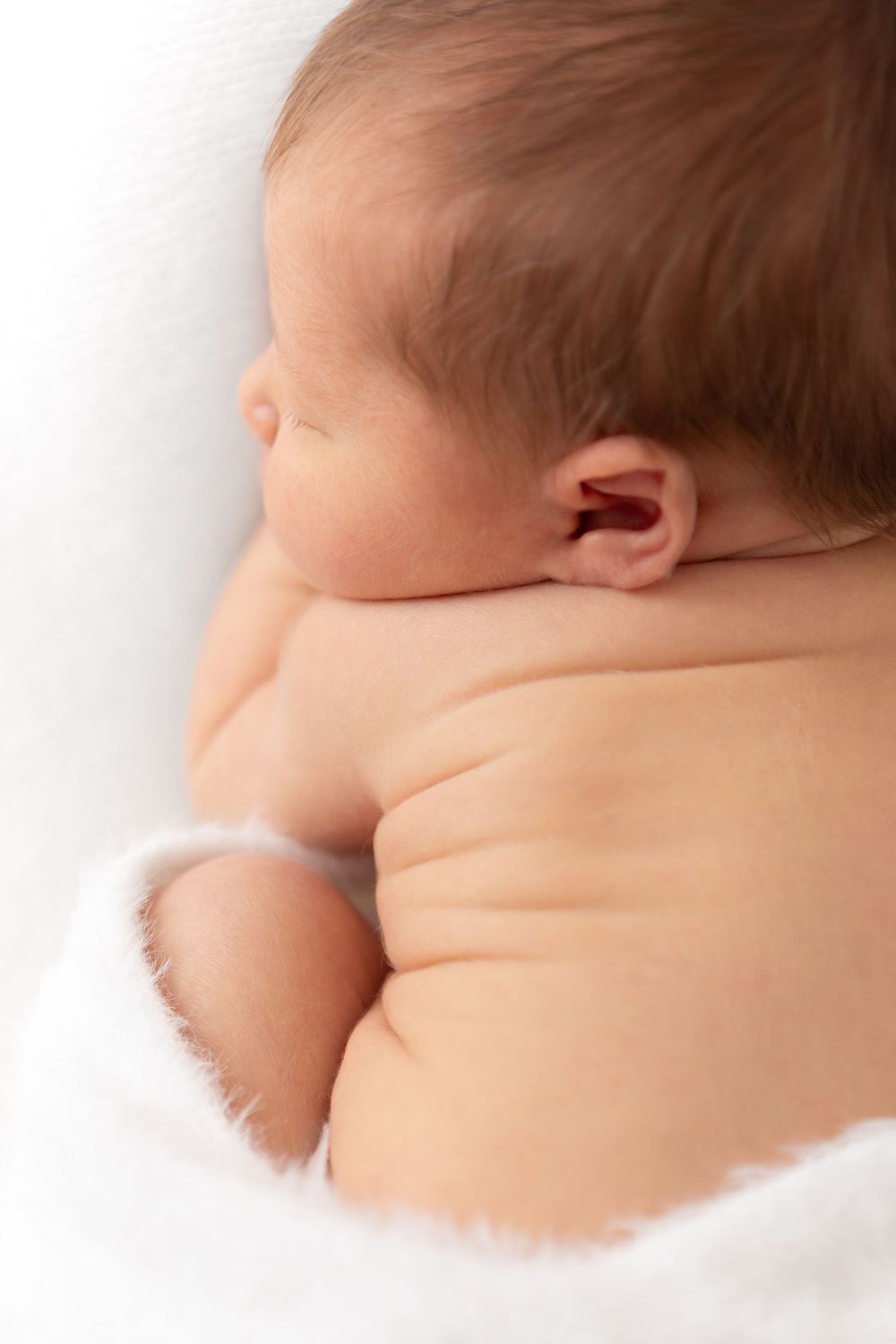 Newborn baby sleeping peacefully on white blanket, showing chubby back and profile view.