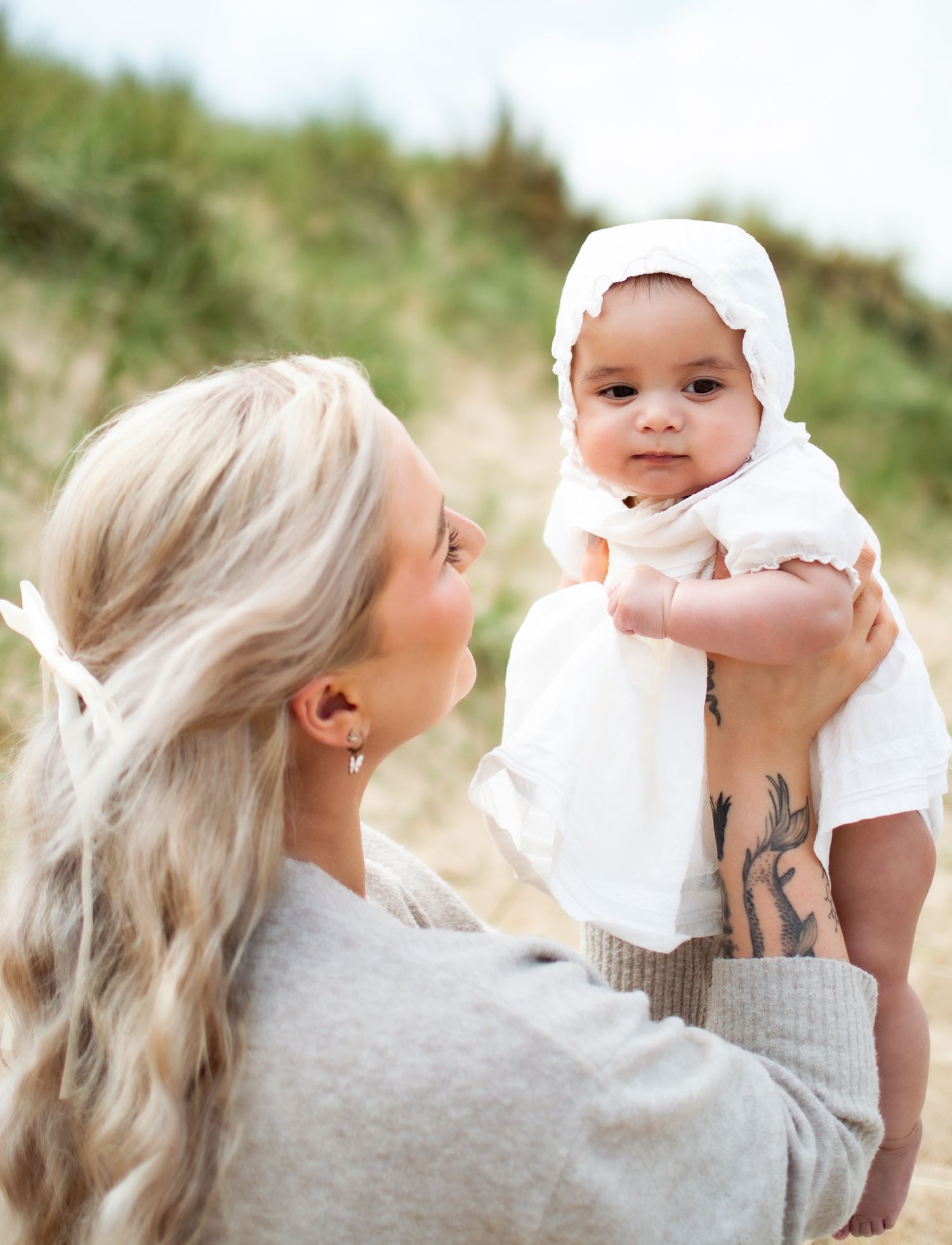 Blonde mother with tattoo lifting smiling baby in white bonnet outdoors on sandy dunes.