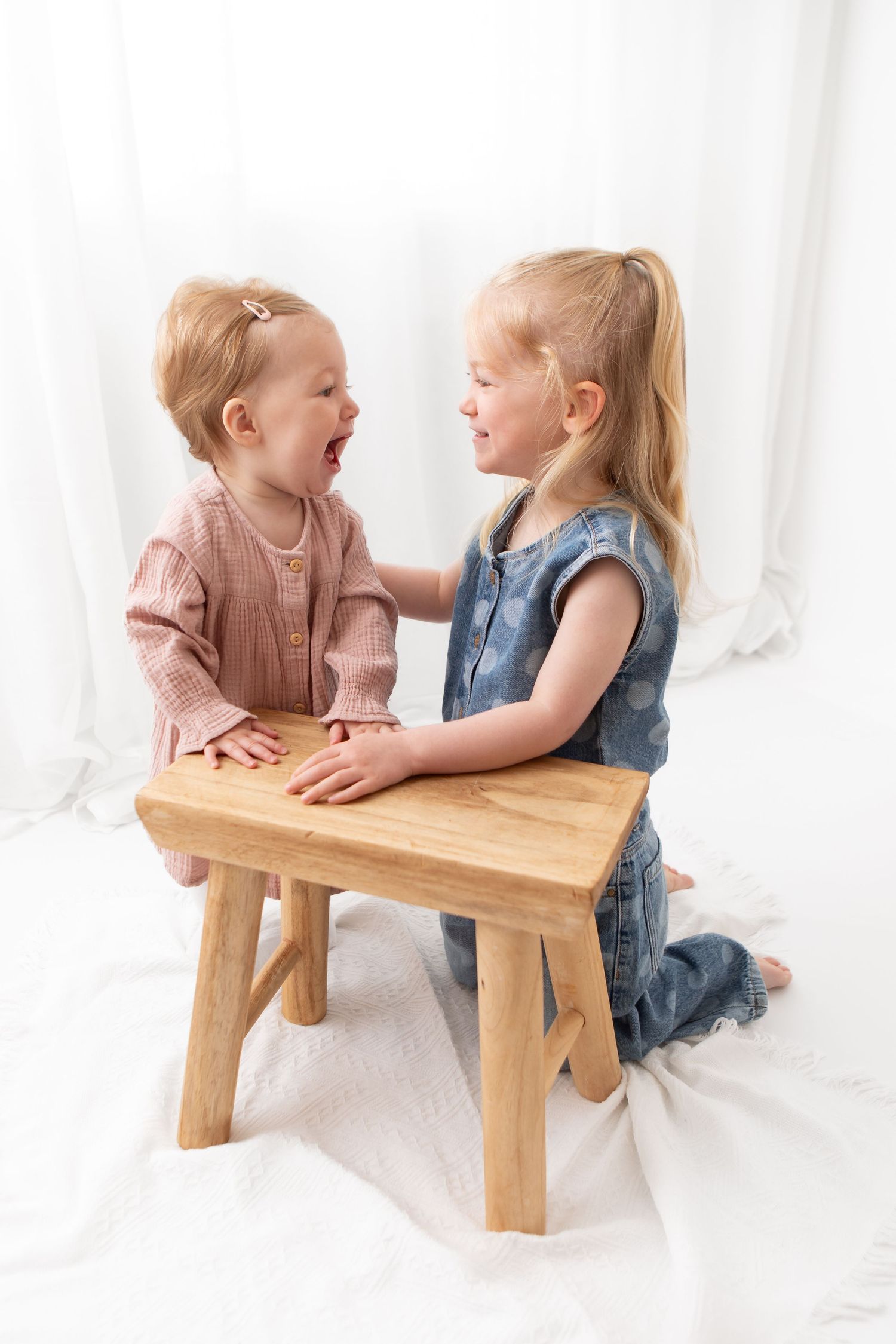 Two young blonde sisters laugh and play together at a small wooden bench in a bright white studio setting.