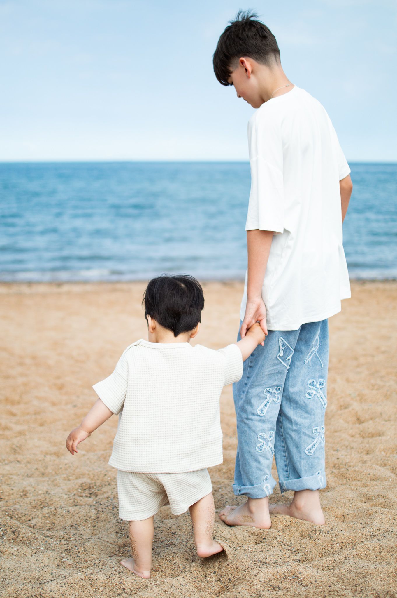 A teenager holds a toddlers hand on a sandy beach, both barefoot wearing white tops and blue bottoms.