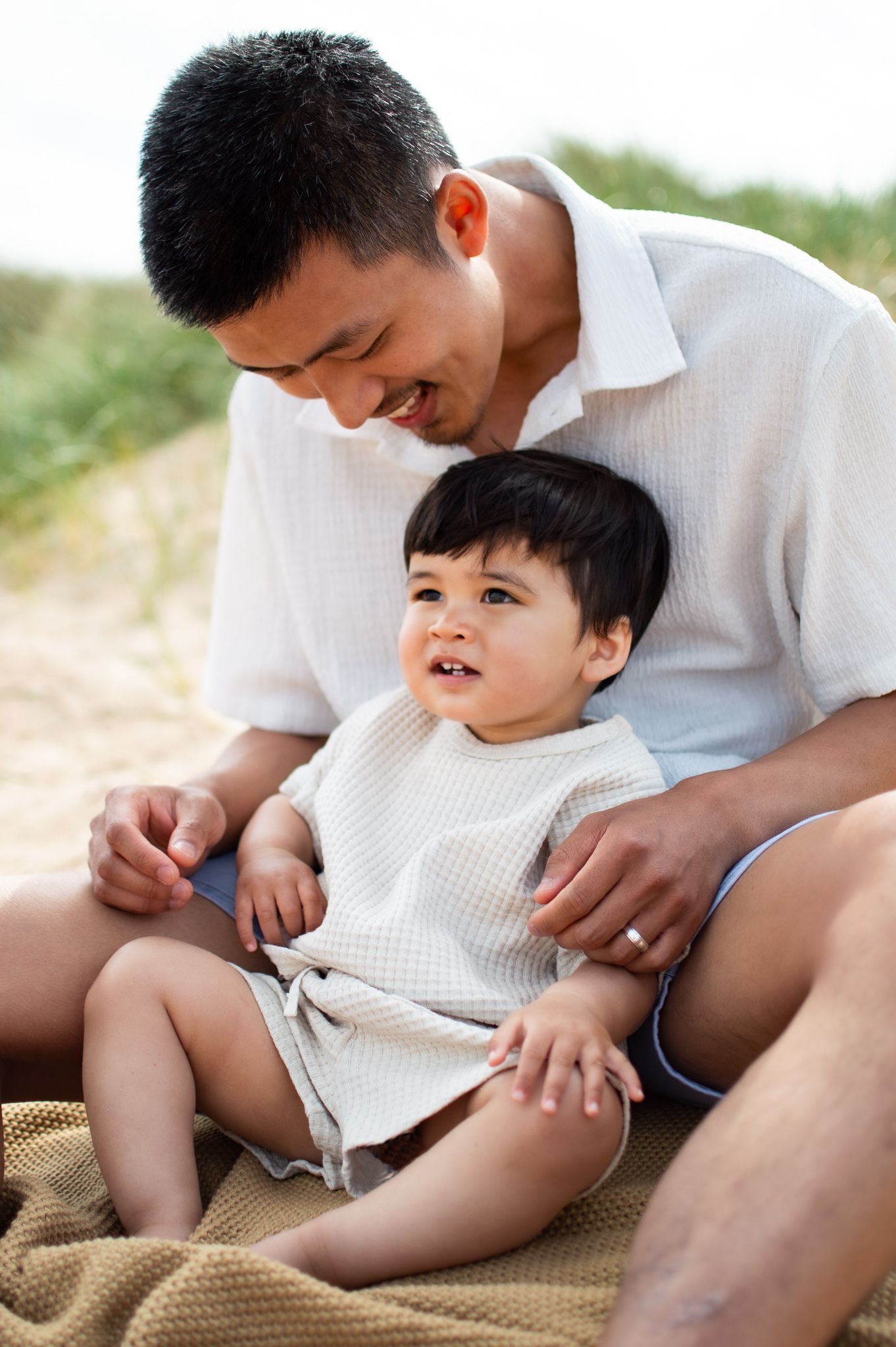 Smiling father sits on beach blanket with happy toddler boy dressed in white waffle outfit.