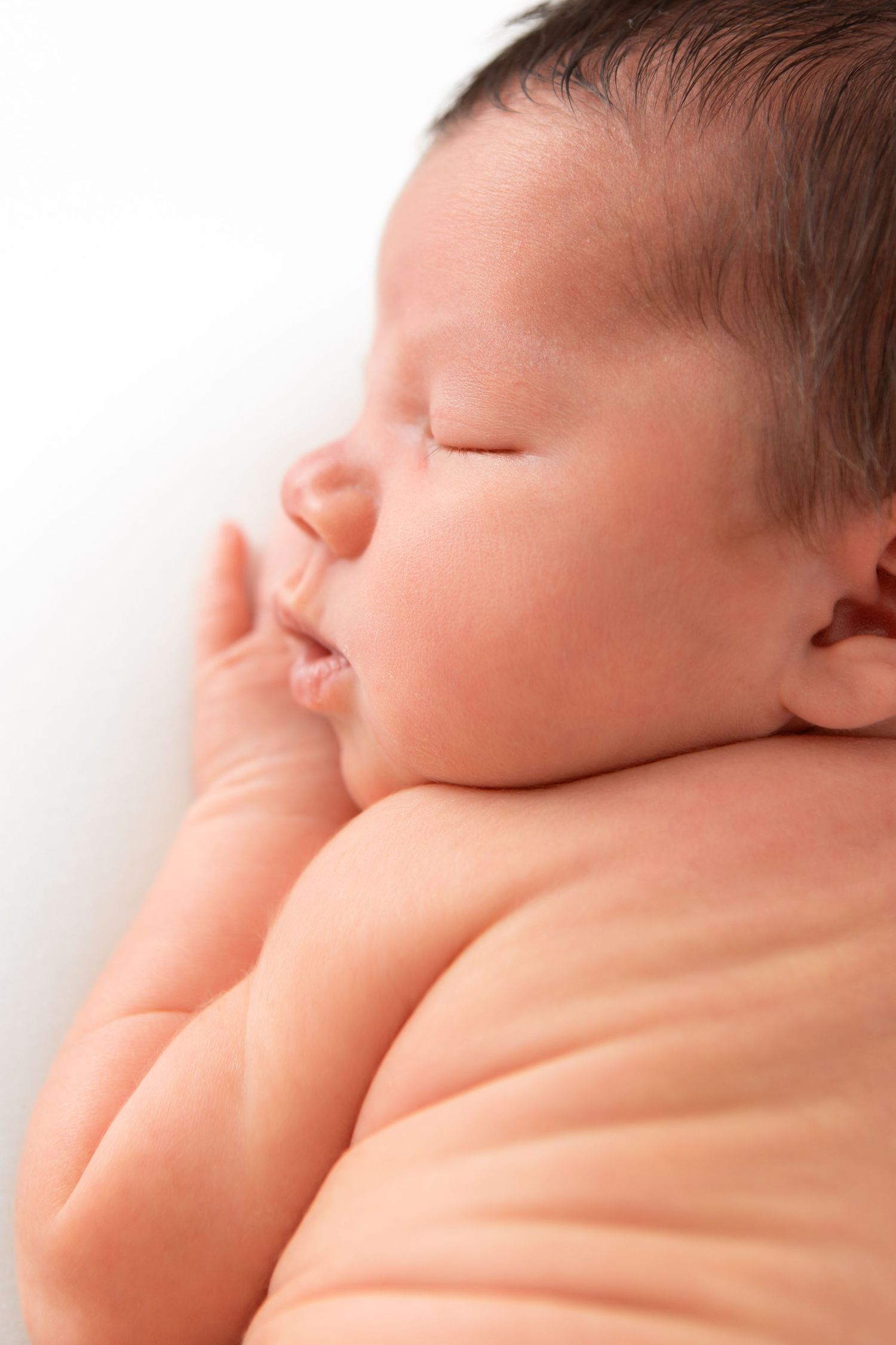 Peaceful newborn baby sleeping soundly with tiny hand near face, resting on white background.
