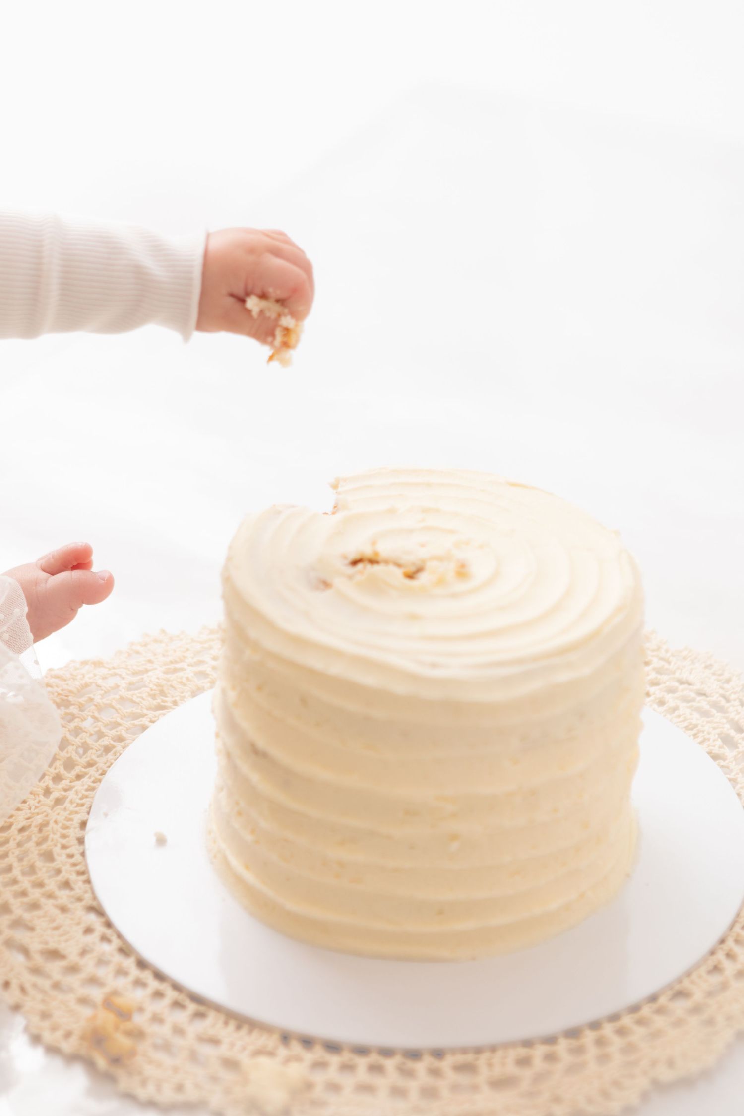 Baby hands reaching into a white frosted smash cake on a lace mat during a first birthday celebration.