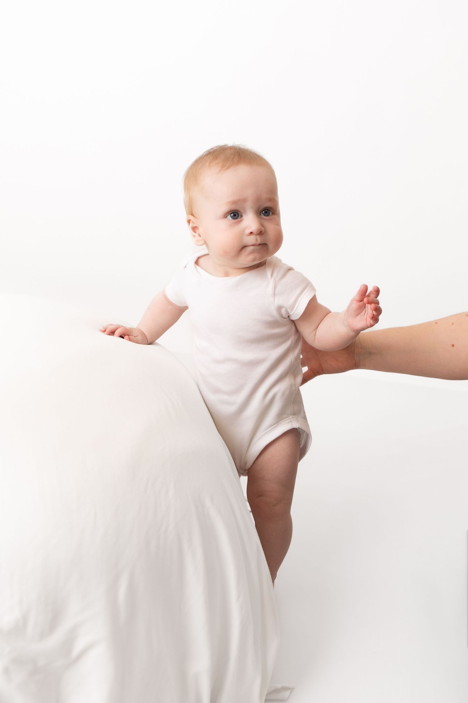 A cute baby in a white onesie stands with adult hand support against a clean white background.