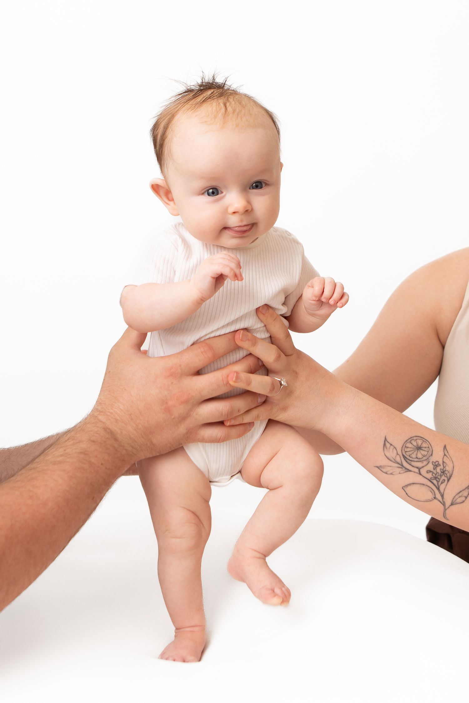 Parents holding a smiling baby in a white onesie against a white background, with a tattooed arm visible.