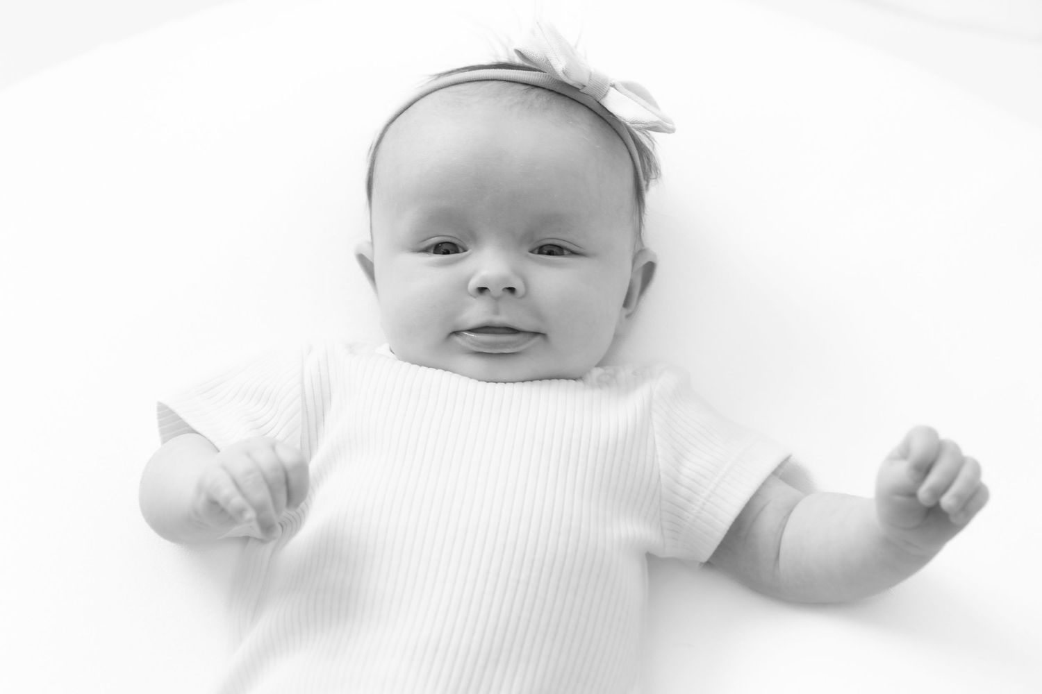 Black and white portrait of a newborn baby in a white onesie with a bow headband lying on a white background.