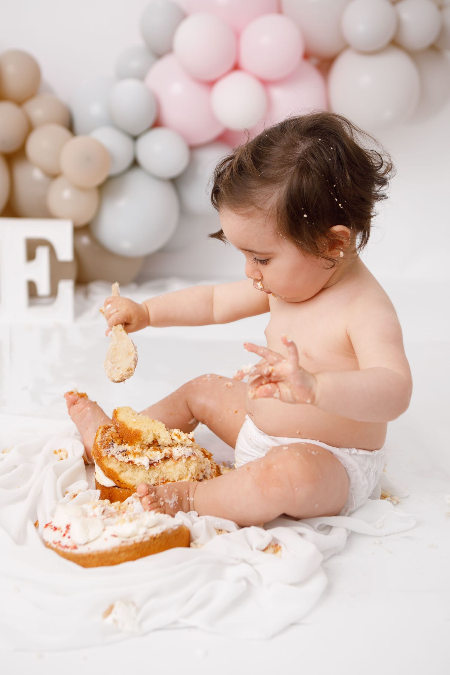 Baby in diaper smashing first birthday cake surrounded by pastel balloons on white background.