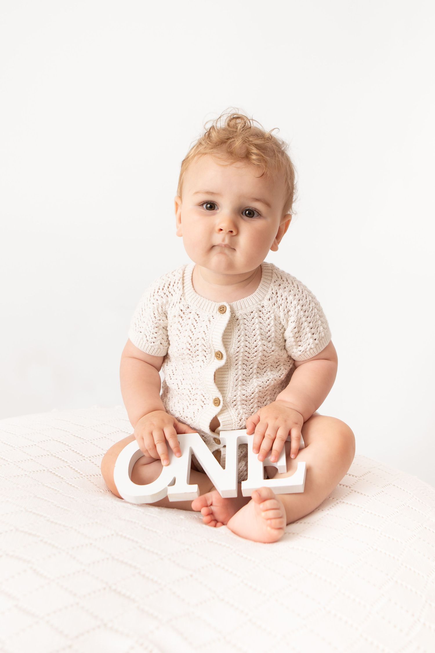 A cute baby in a cream knit romper sits on a white surface holding a wooden ONE sign for their first birthday.