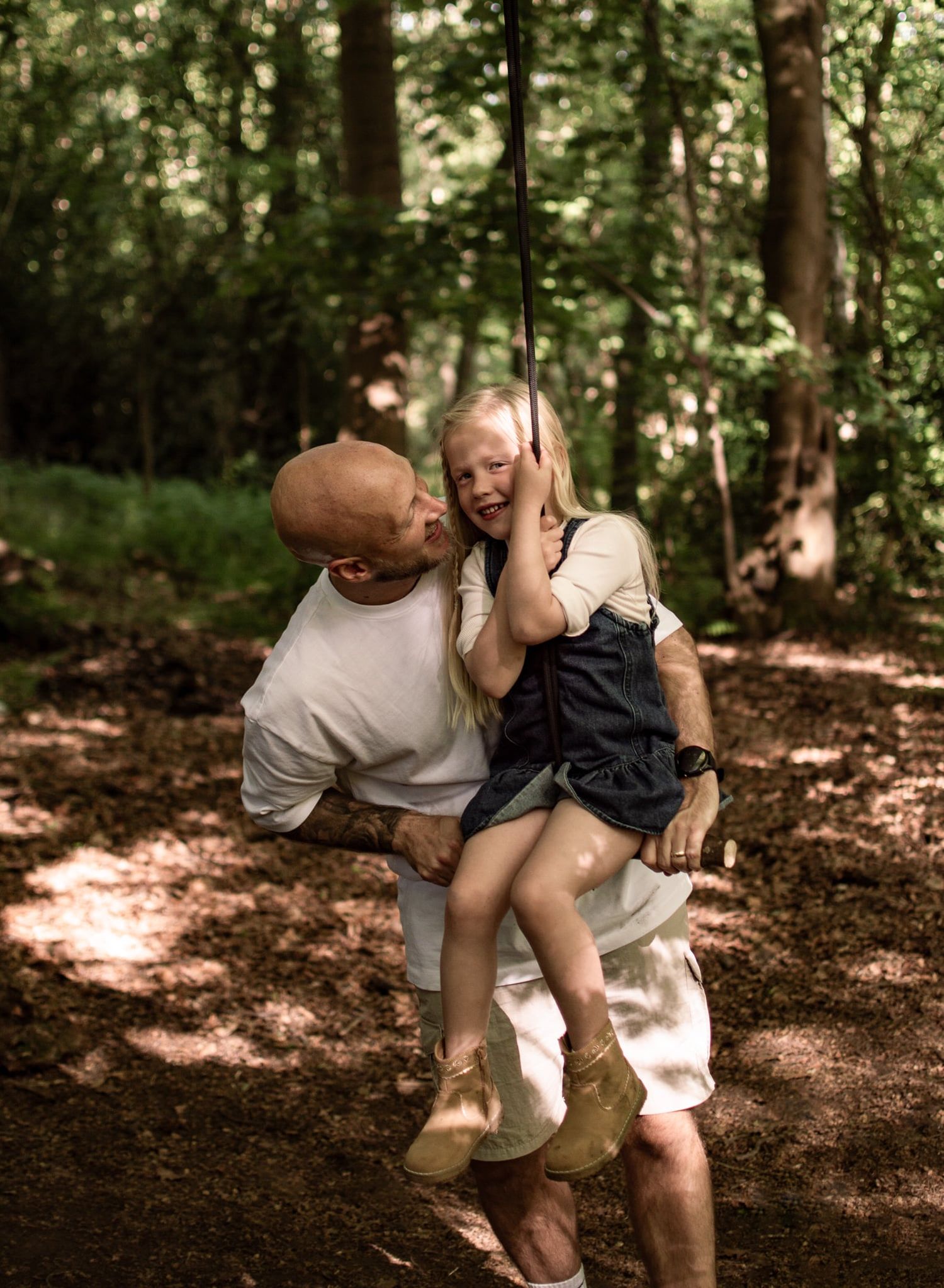 Bald man kissing cheek of laughing young girl on rope swing in shaded woodland setting.