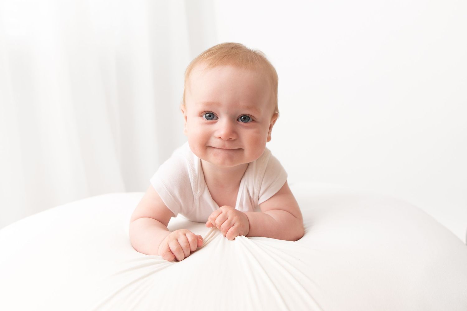 Smiling baby in white onesie doing tummy time on a white surface in a bright, airy room.